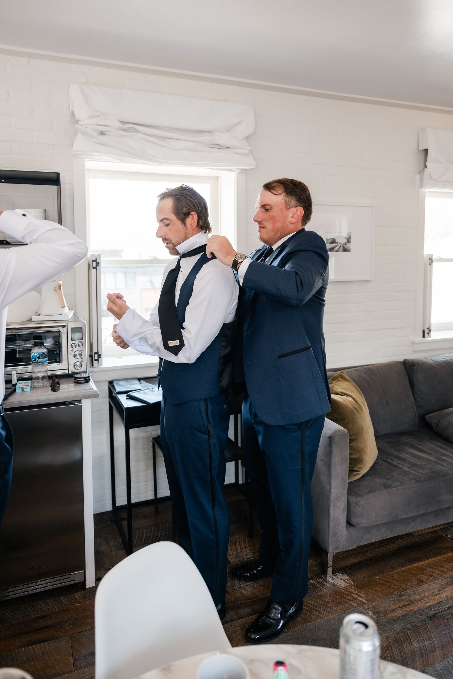Wedding party members helping with formal attire preparations in a bright room.