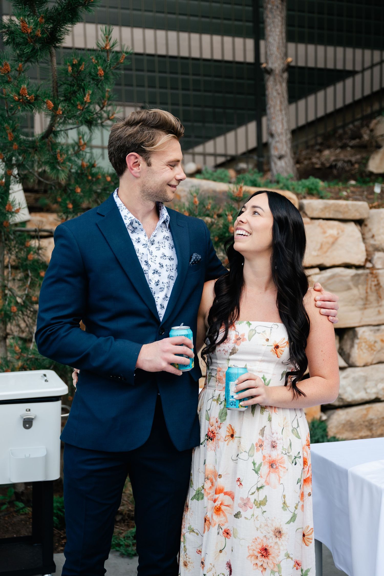 Candid reception moment of couple sharing drinks and laughter by stone wall garden.