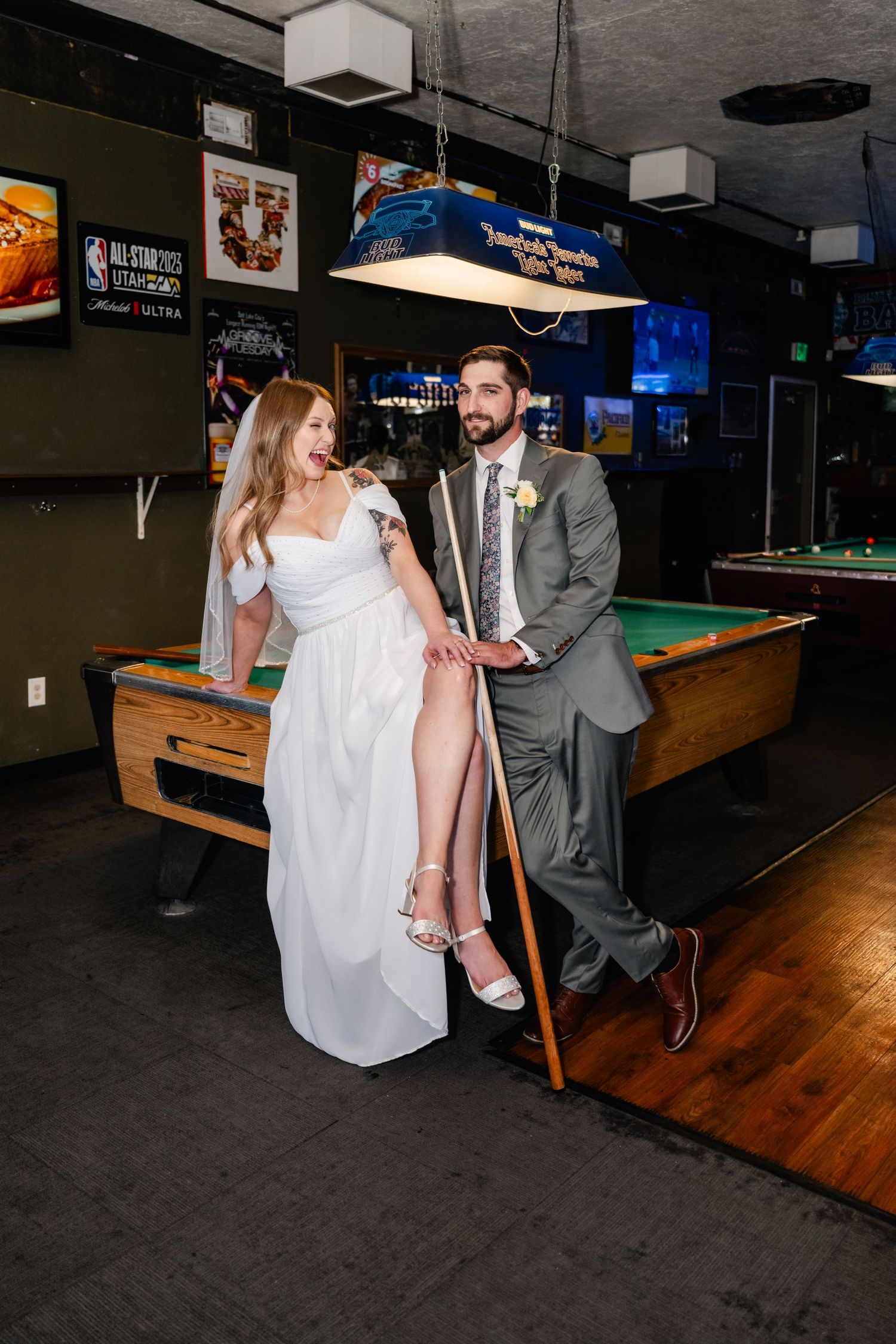 A couple in wedding attire poses playfully near a pool table in a bar setting.