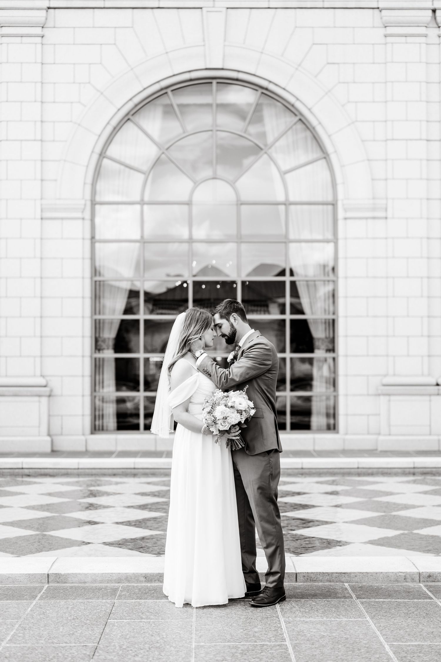 A romantic wedding couple embraces on a grand checkered courtyard with arched windows in the background.