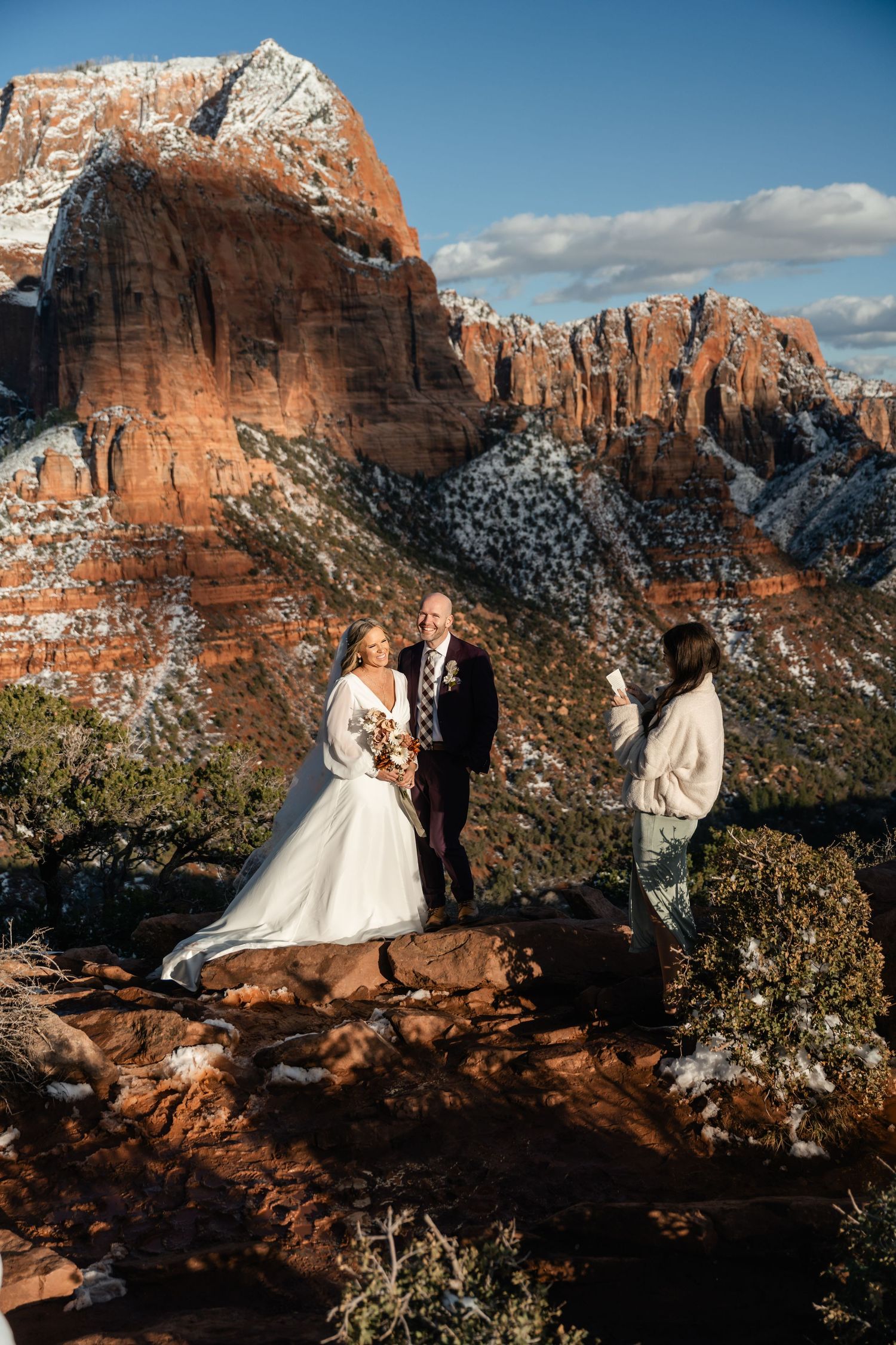 Wedding ceremony taking place on red rock cliffs with snow-capped mountains in background.