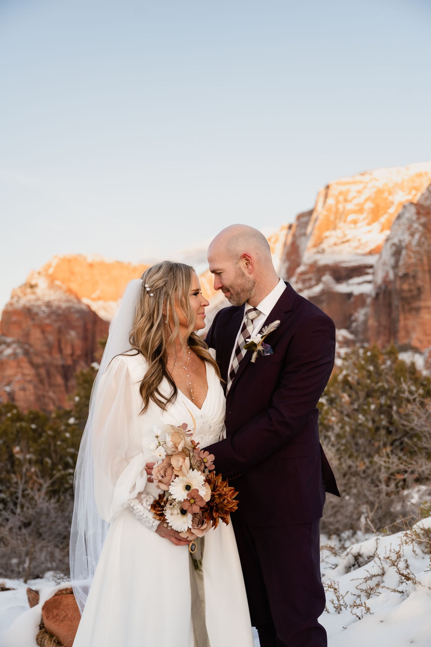 Wedding couple sharing intimate moment against backdrop of snow-dusted red rock cliffs at sunset.