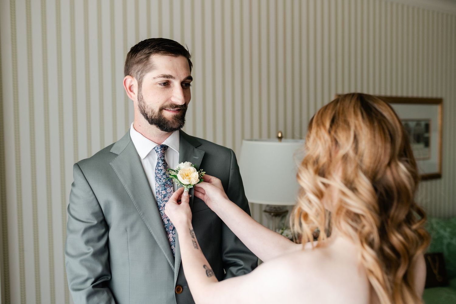 Someone pins a white boutonniere flower onto a gray suit jacket lapel during wedding preparations.