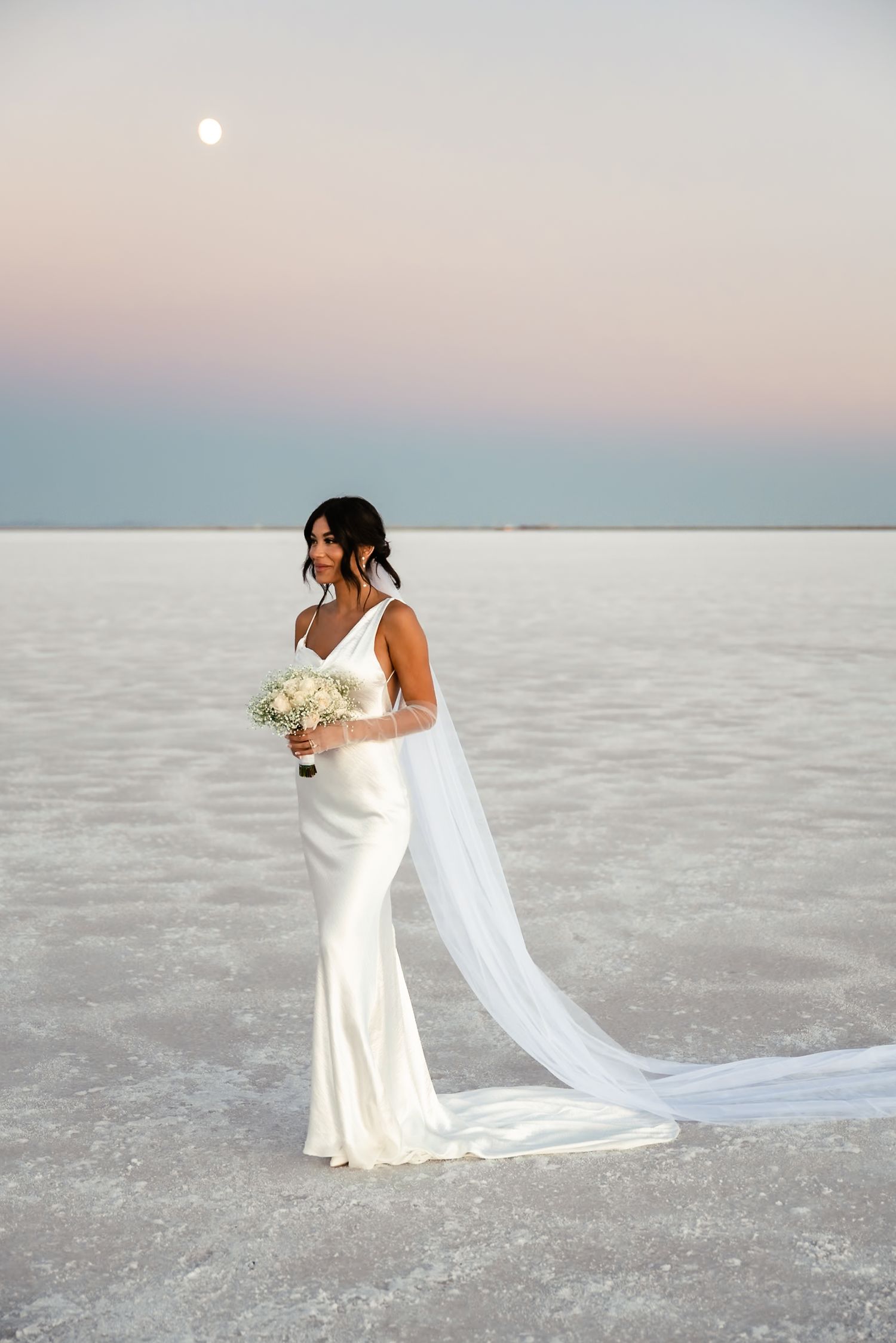 A flowing white wedding dress and veil catch the breeze against a serene salt flat backdrop.