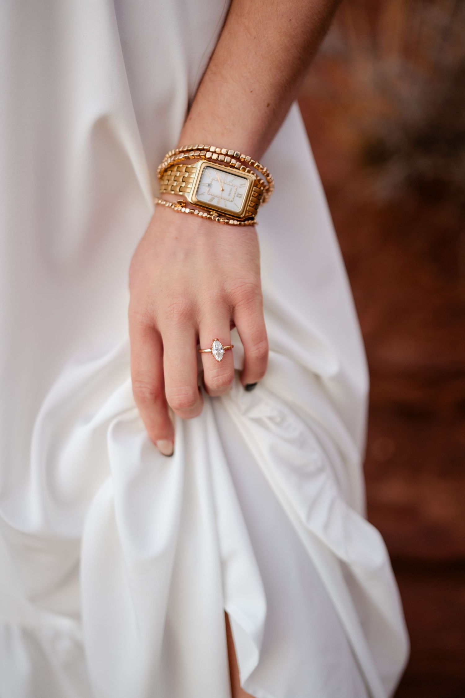 Close-up of a hand wearing an elegant gold watch and diamond engagement ring against white fabric.