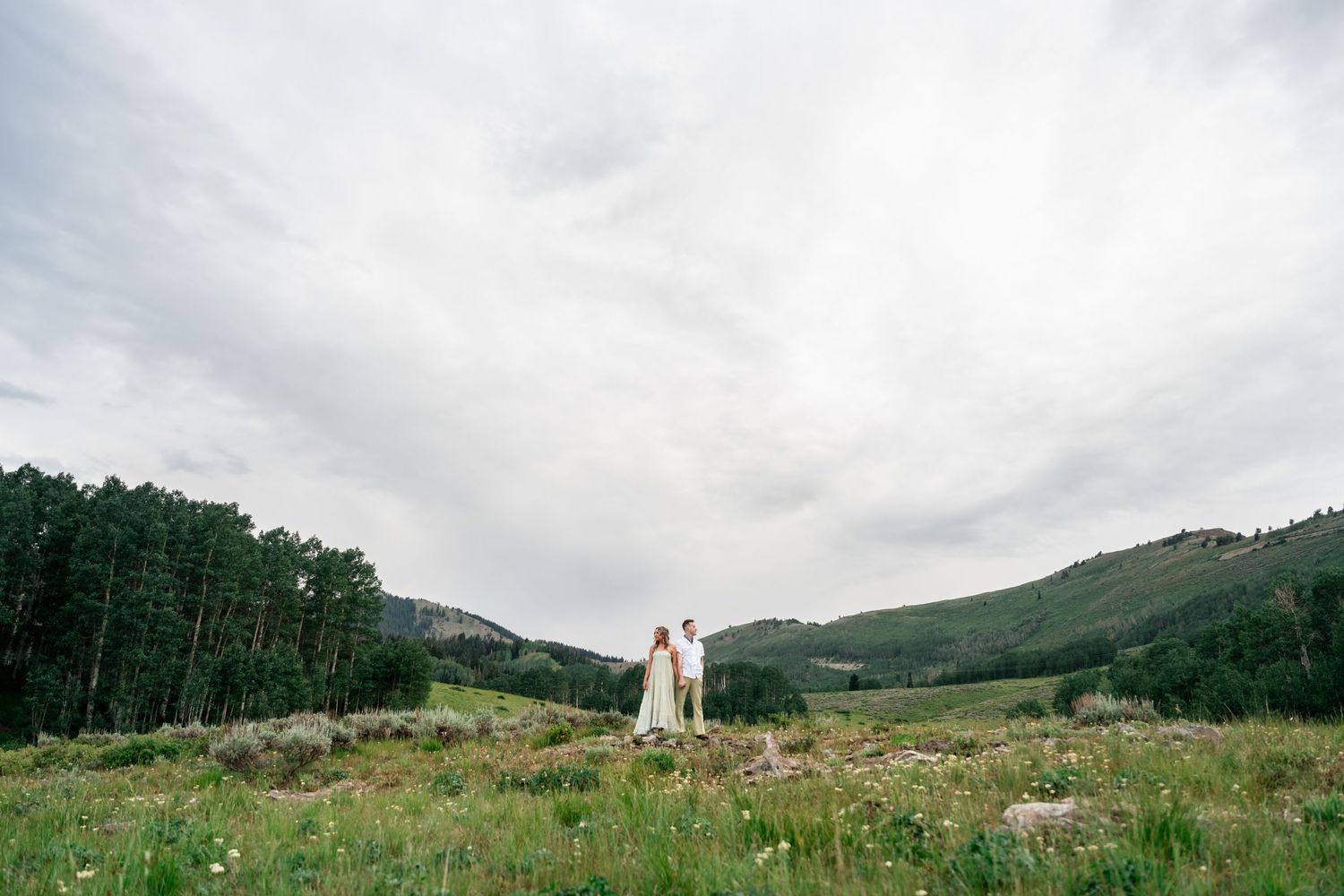 A couple stands in a scenic mountain meadow surrounded by evergreen forests and rolling hills under cloudy skies.