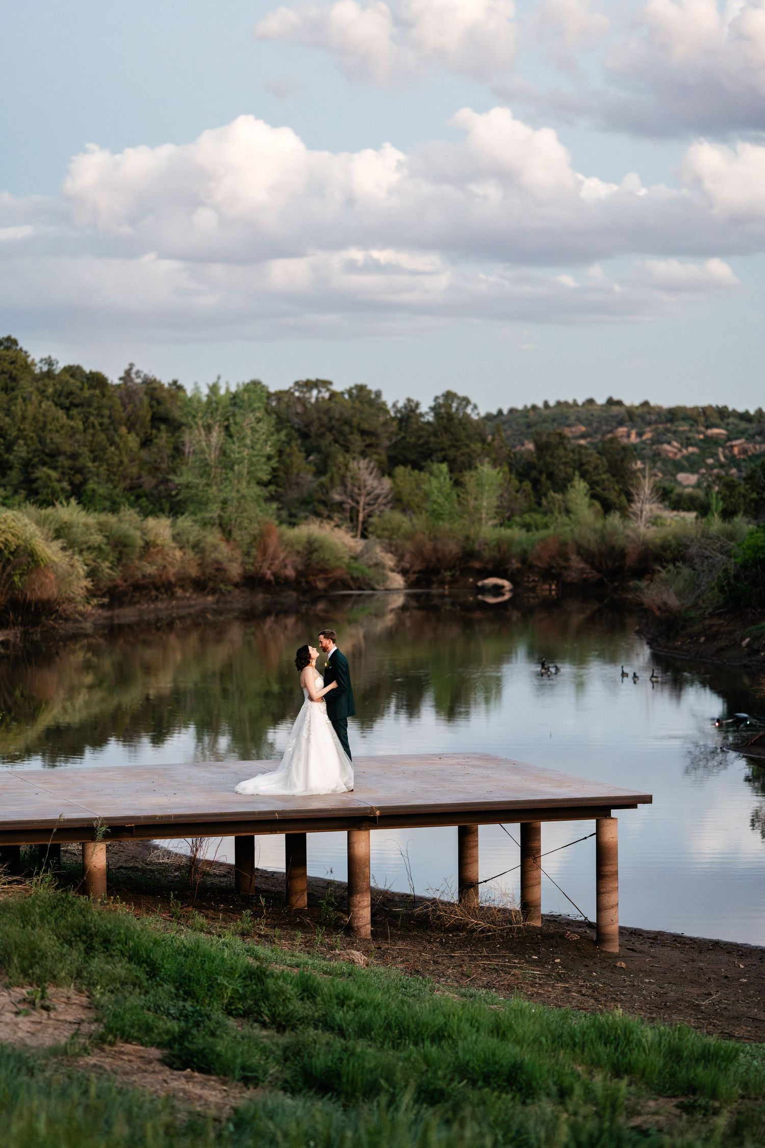 Wedding couple standing on wooden pier overlooking serene lake with cloudy sky.