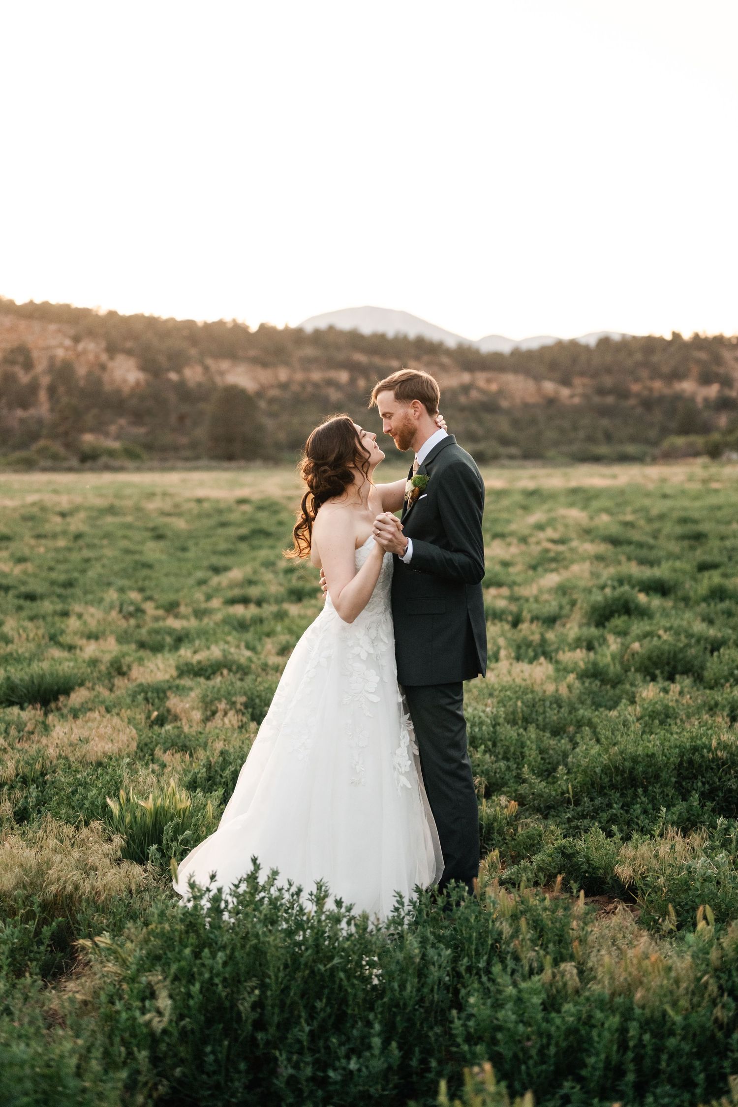 A couple shares an intimate moment in formal wear against a backdrop of green hills at sunset.