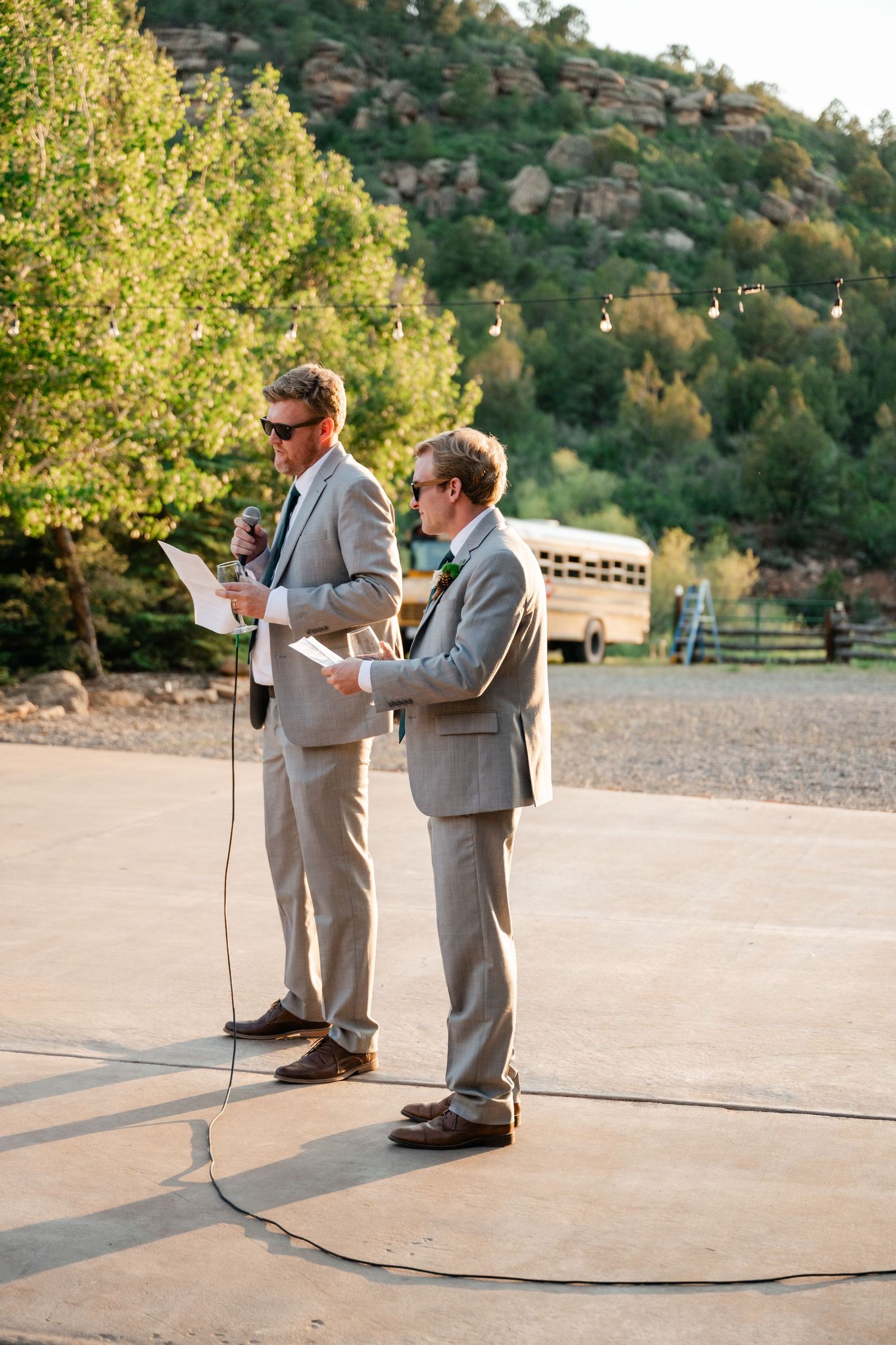 Two people in gray suits reading from papers at an outdoor ceremony with a hillside backdrop.