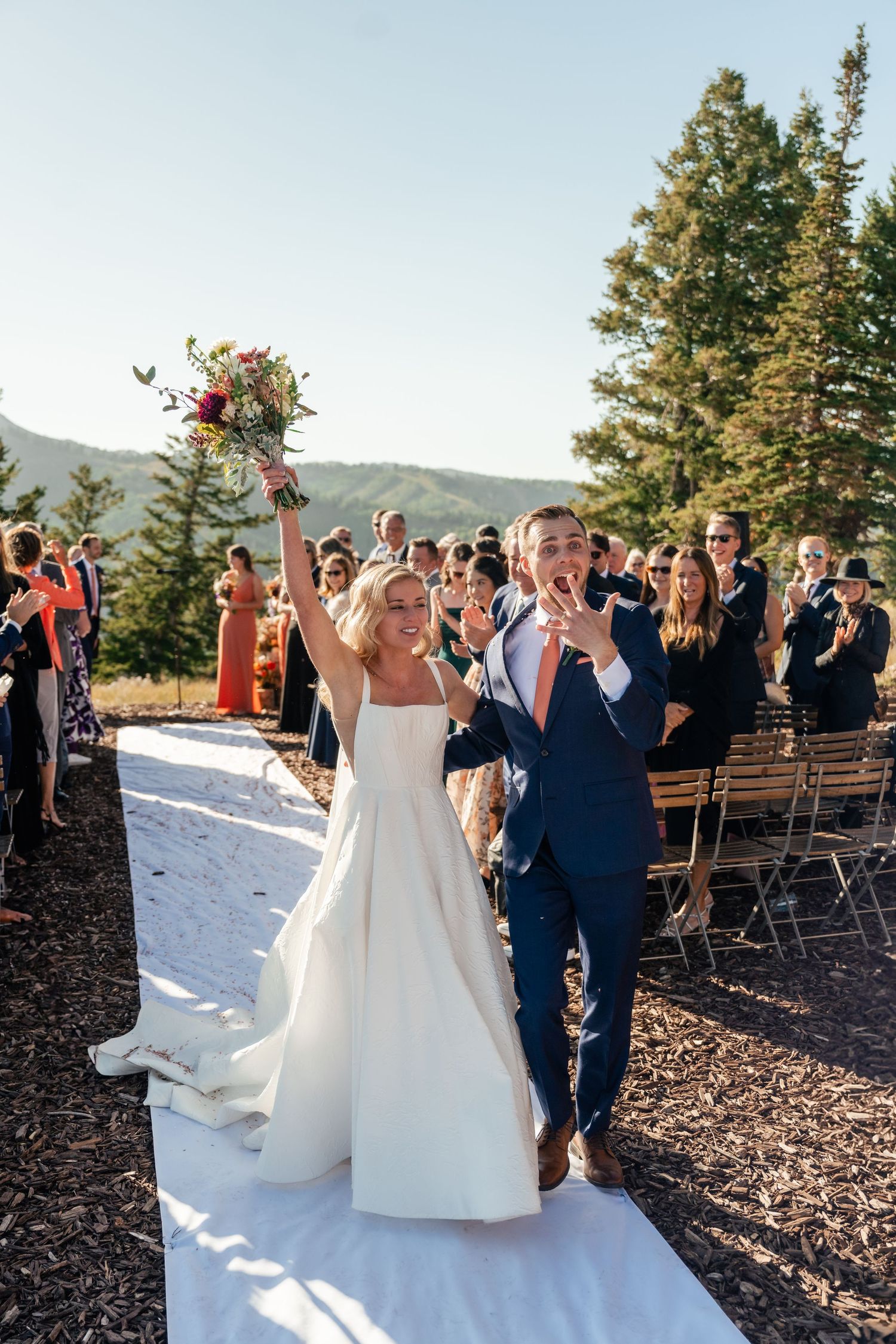 Newlyweds celebrate with raised arms during their mountaintop wedding ceremony as guests cheer and applaud. Park City Wedding Photographer.