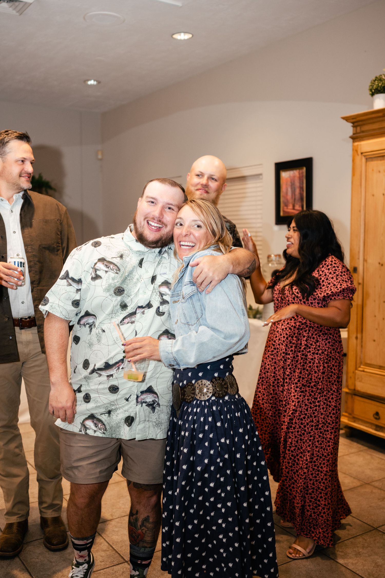 Party guests enjoying drinks and laughter at an indoor celebration with wooden furniture and warm lighting.