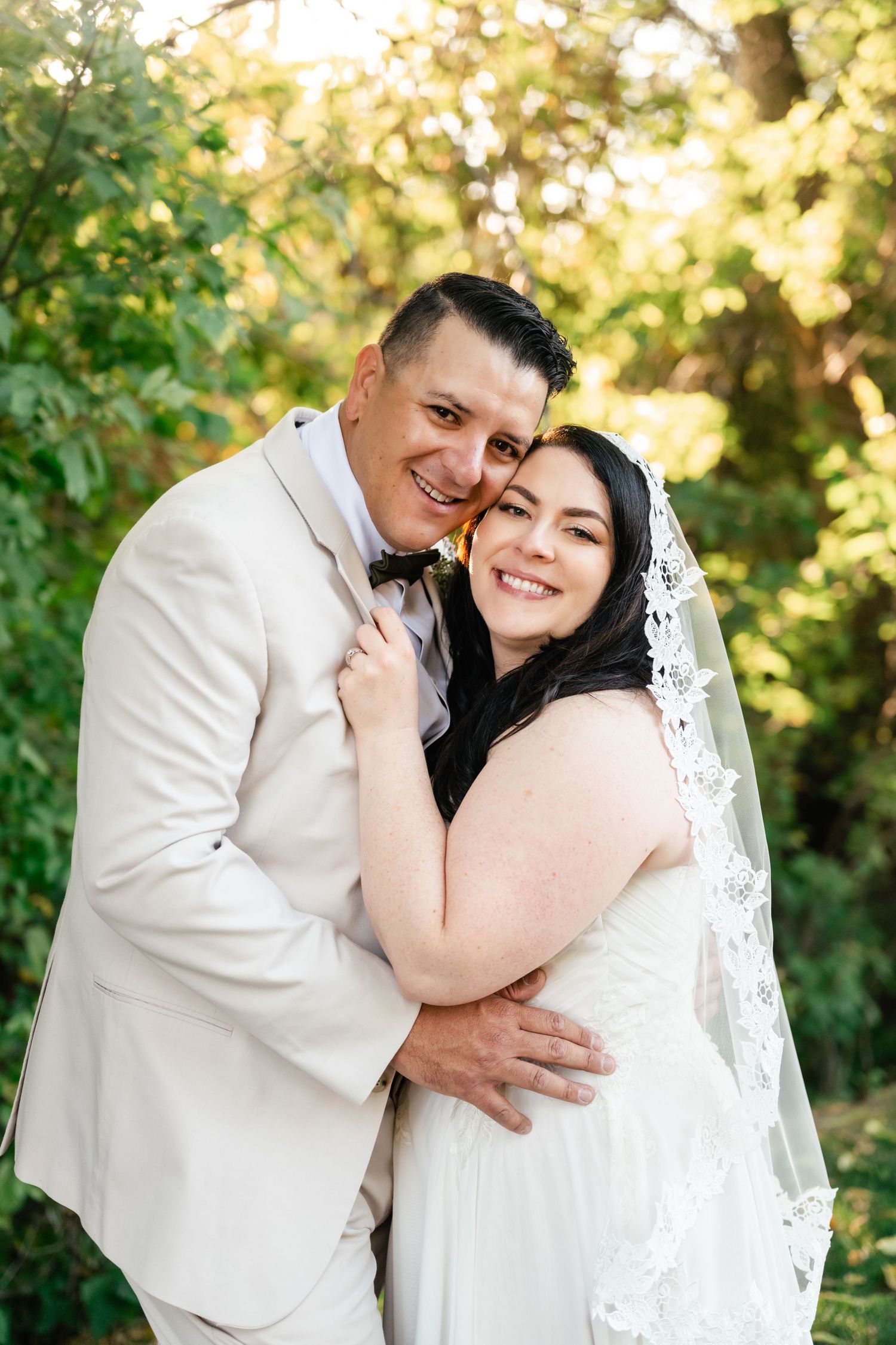 A joyful couple embraces outdoors in wedding attire with lush greenery and sunlit foliage in the background.