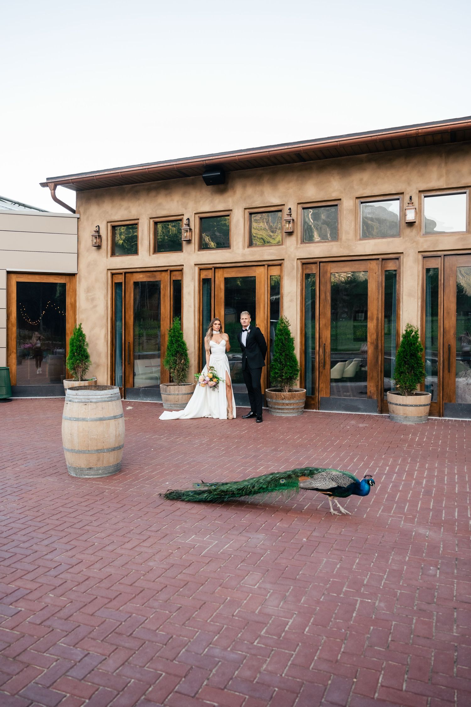 Wedding couple stands in front of rustic venue with large wooden barrel decor on brick patio in garden setting.
