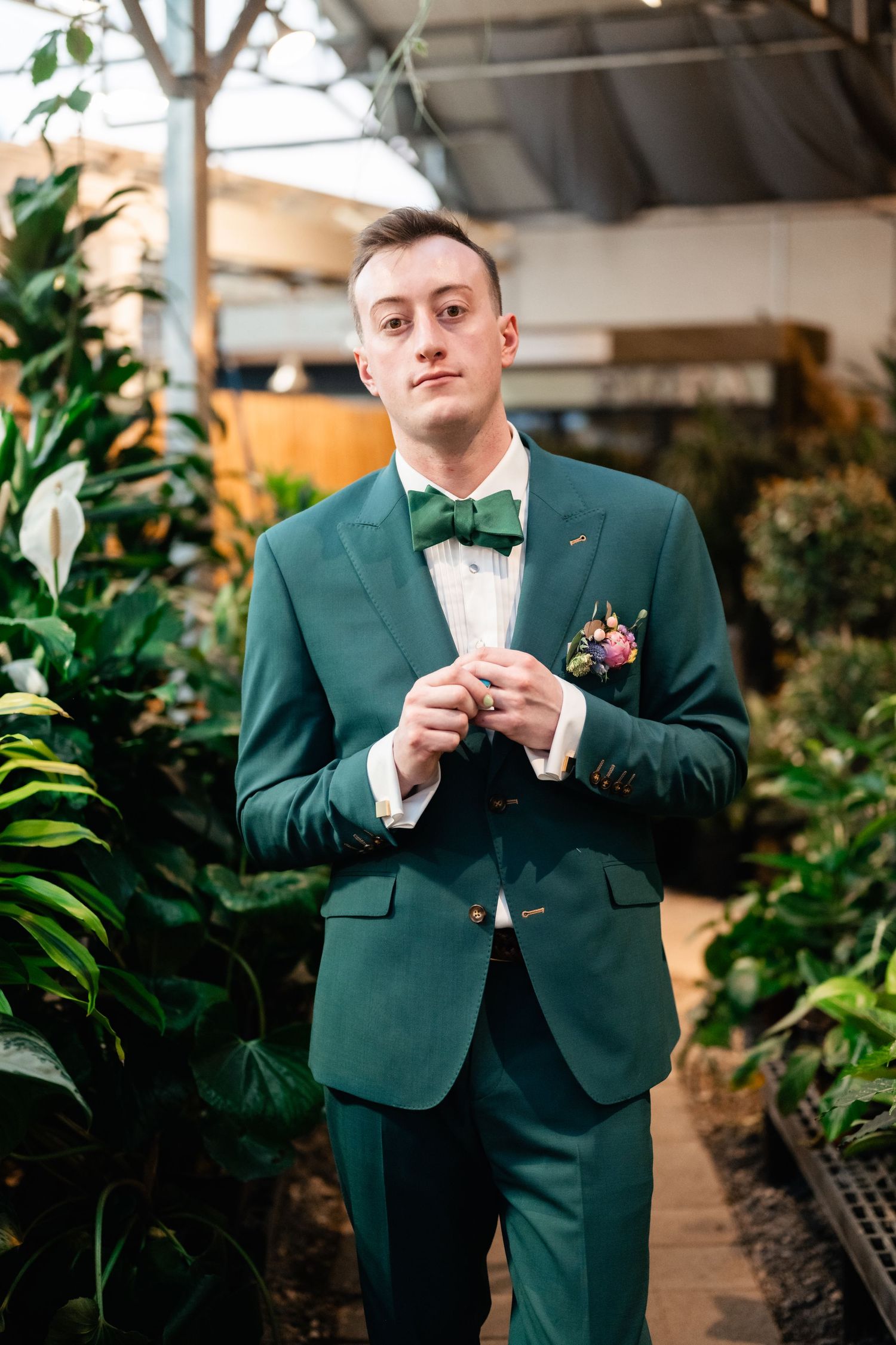 A person in an emerald green tuxedo with bow tie stands in a greenhouse surrounded by lush plants and greenery.