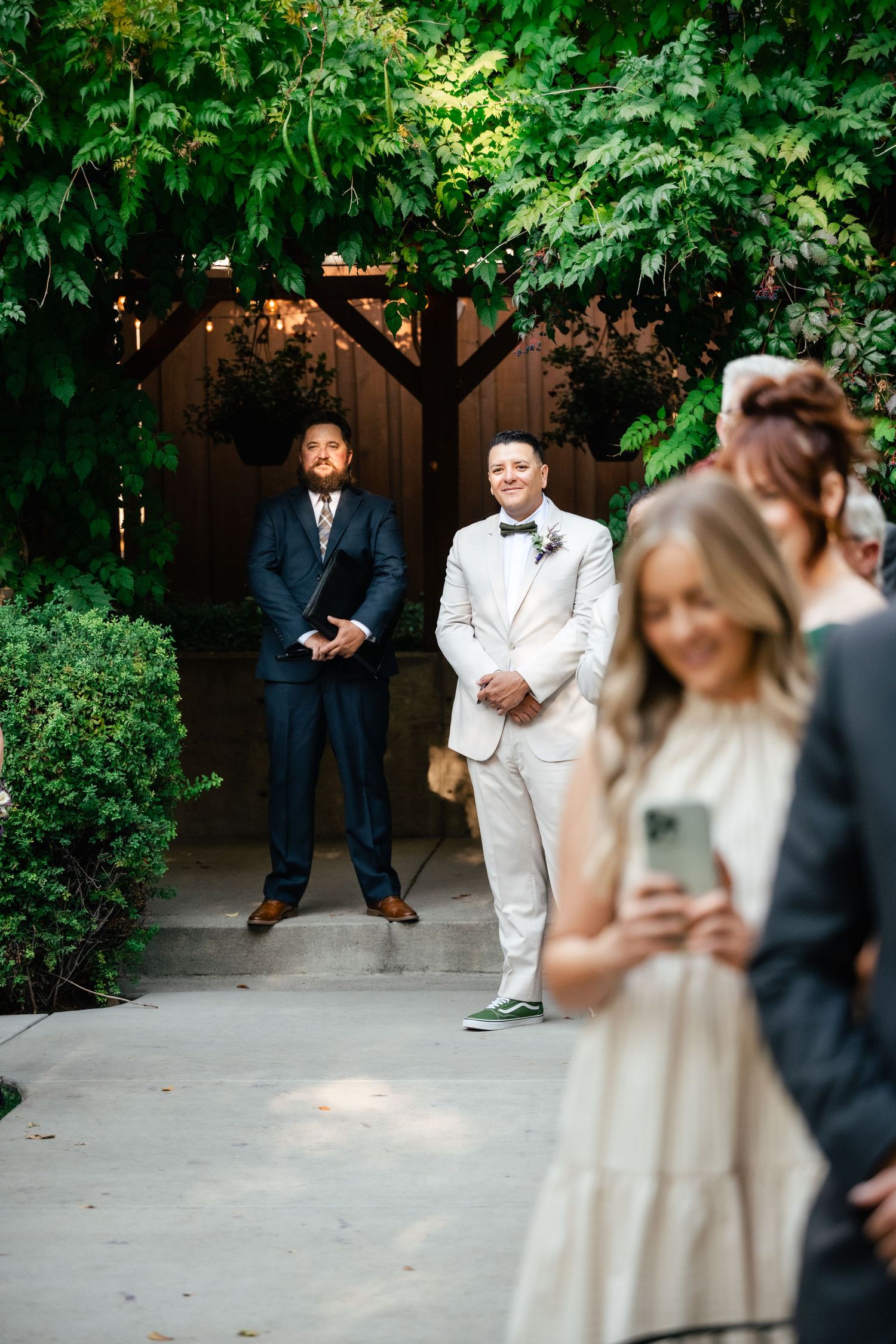 Two wedding guests stand in a garden pathway while others socialize in the foreground at an outdoor ceremony.