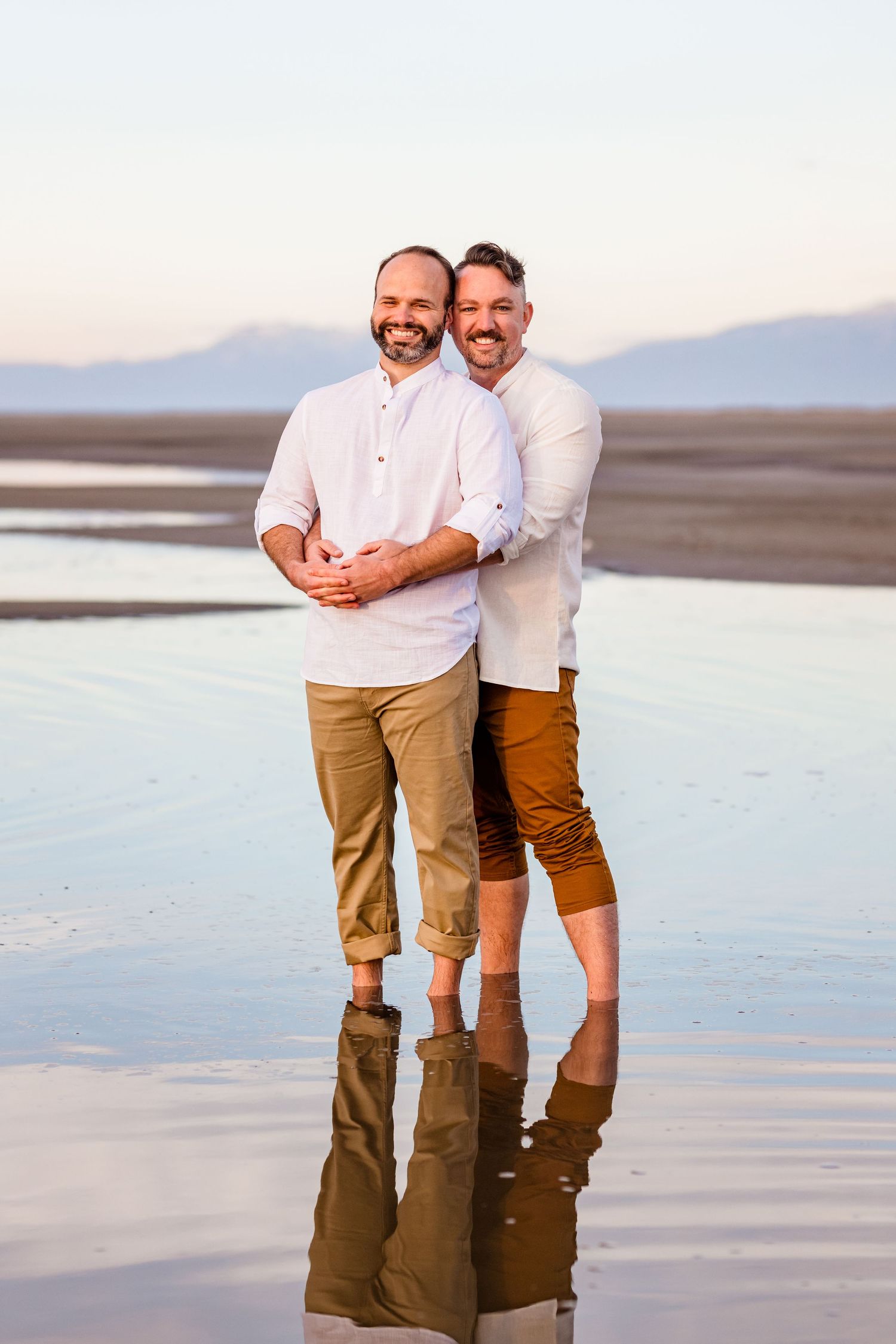 Couple embracing on reflective salt flats during sunset with mountains in background.
