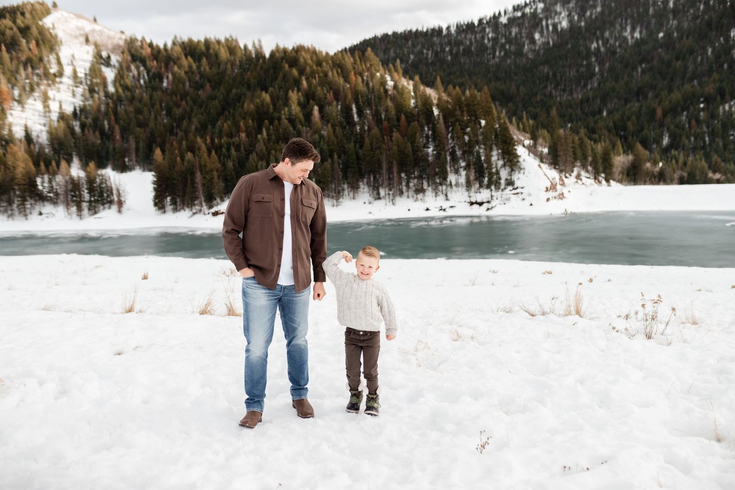 A couple walks together through a snowy winter landscape with evergreen trees and mountains in the background.