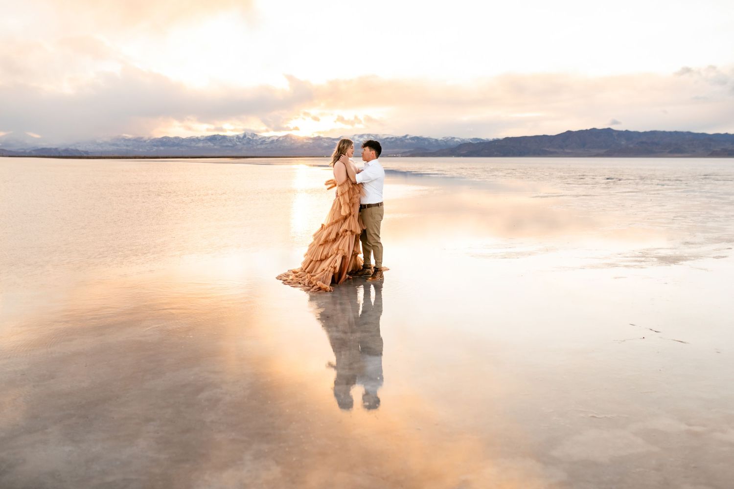 Utah Salt Flats Photos. A couple stands in shallow reflective water at sunset with mountains in the background and golden light all around.