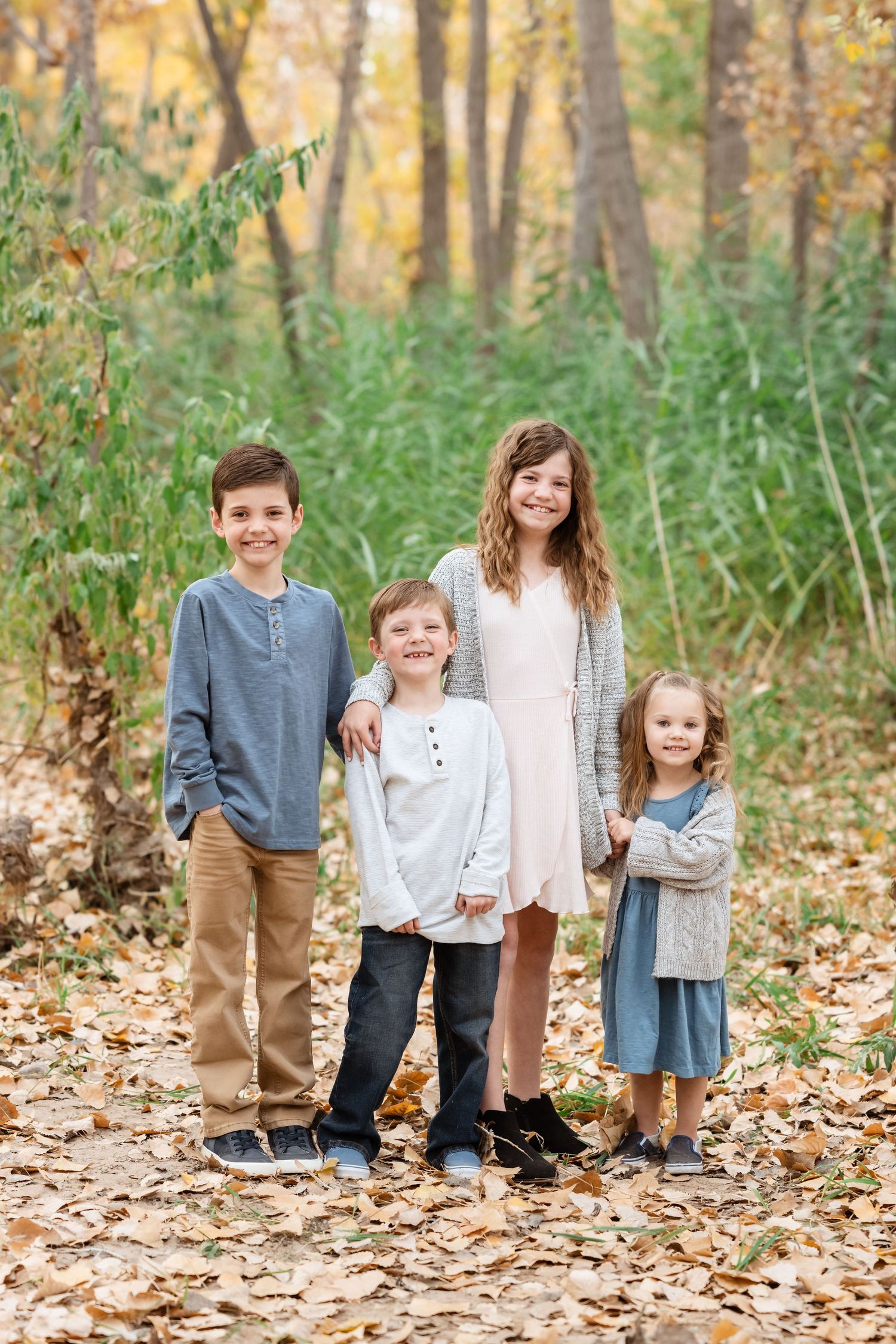 Four children stand together on fallen autumn leaves in a wooded area wearing coordinated casual outfits.