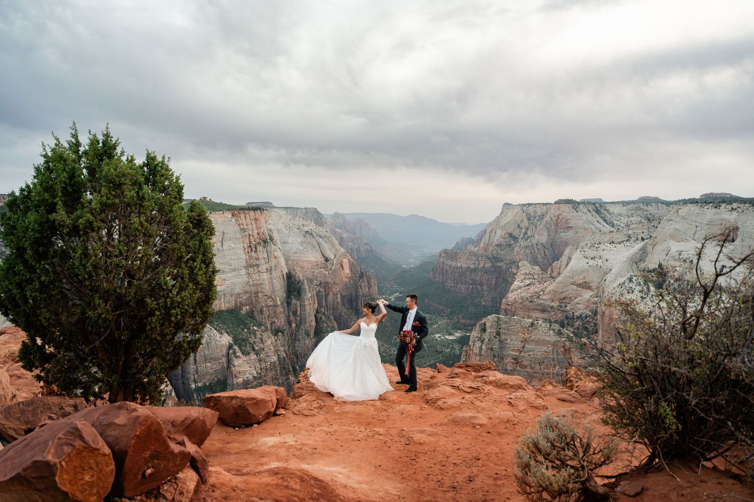 Two people in wedding attire stand on red rocks overlooking the dramatic canyon landscape at Zion National Park.