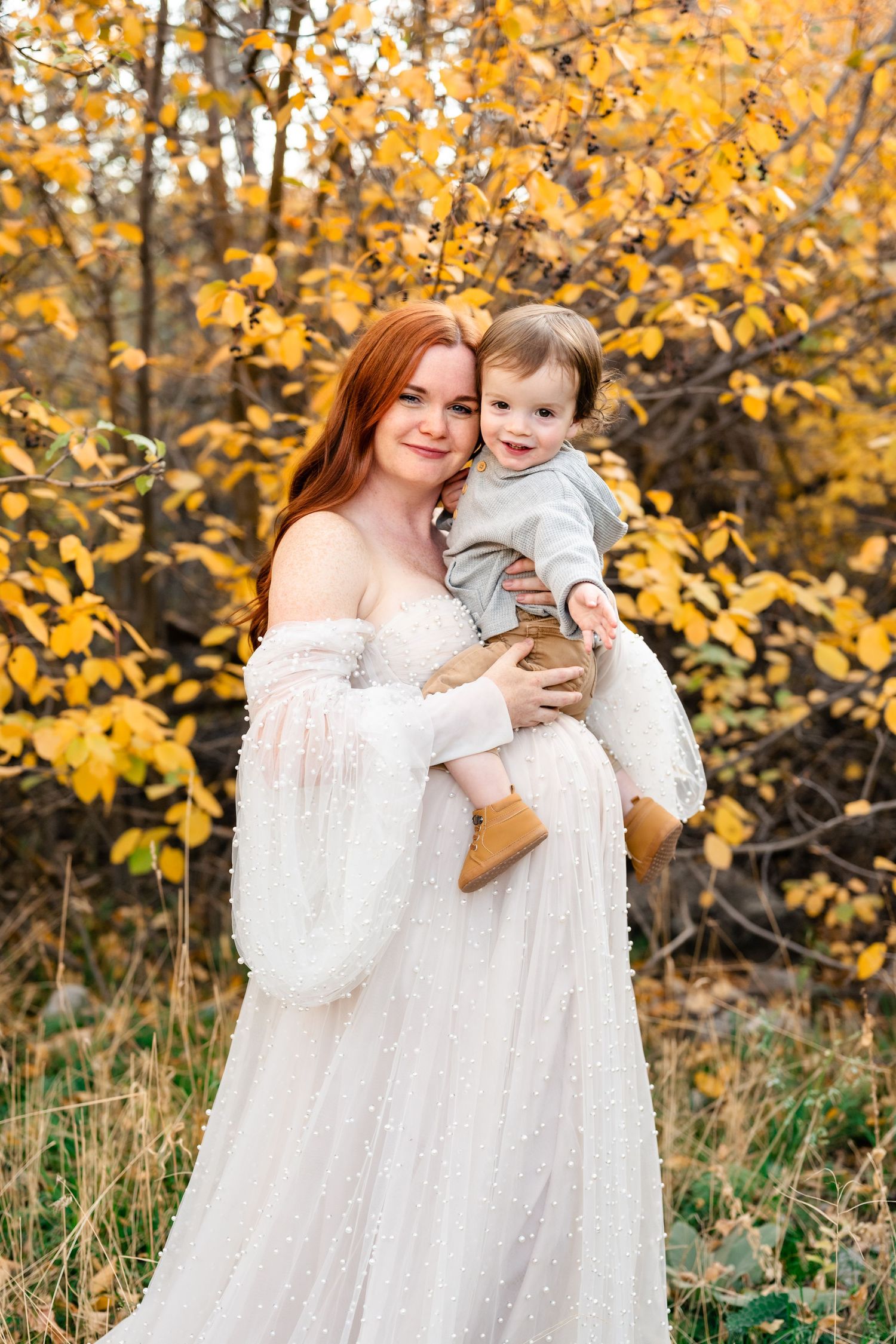 Mother and child in matching white dresses share joyful moment against backdrop of golden fall leaves.