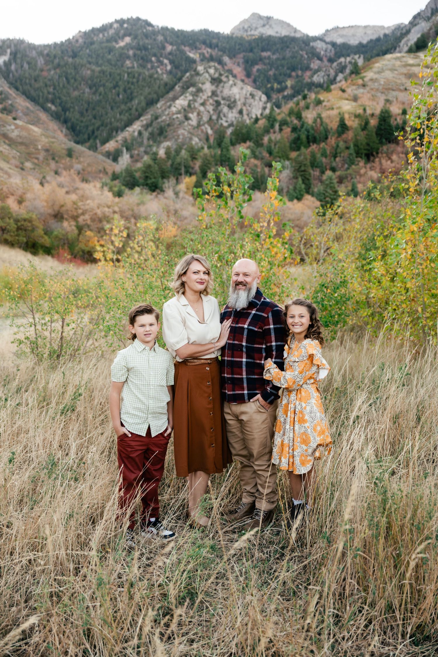 A group poses together in tall autumn grass with mountains and fall foliage creating a scenic outdoor backdrop.