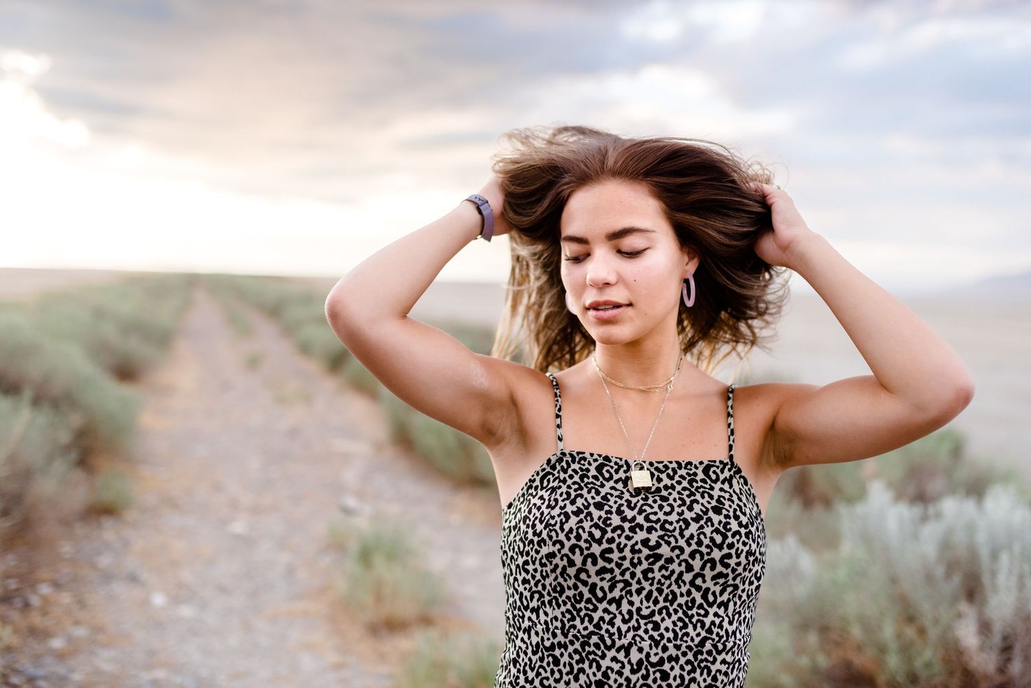 A woman in a leopard print dress stands on a dirt path with windswept hair against a desert landscape at sunset.