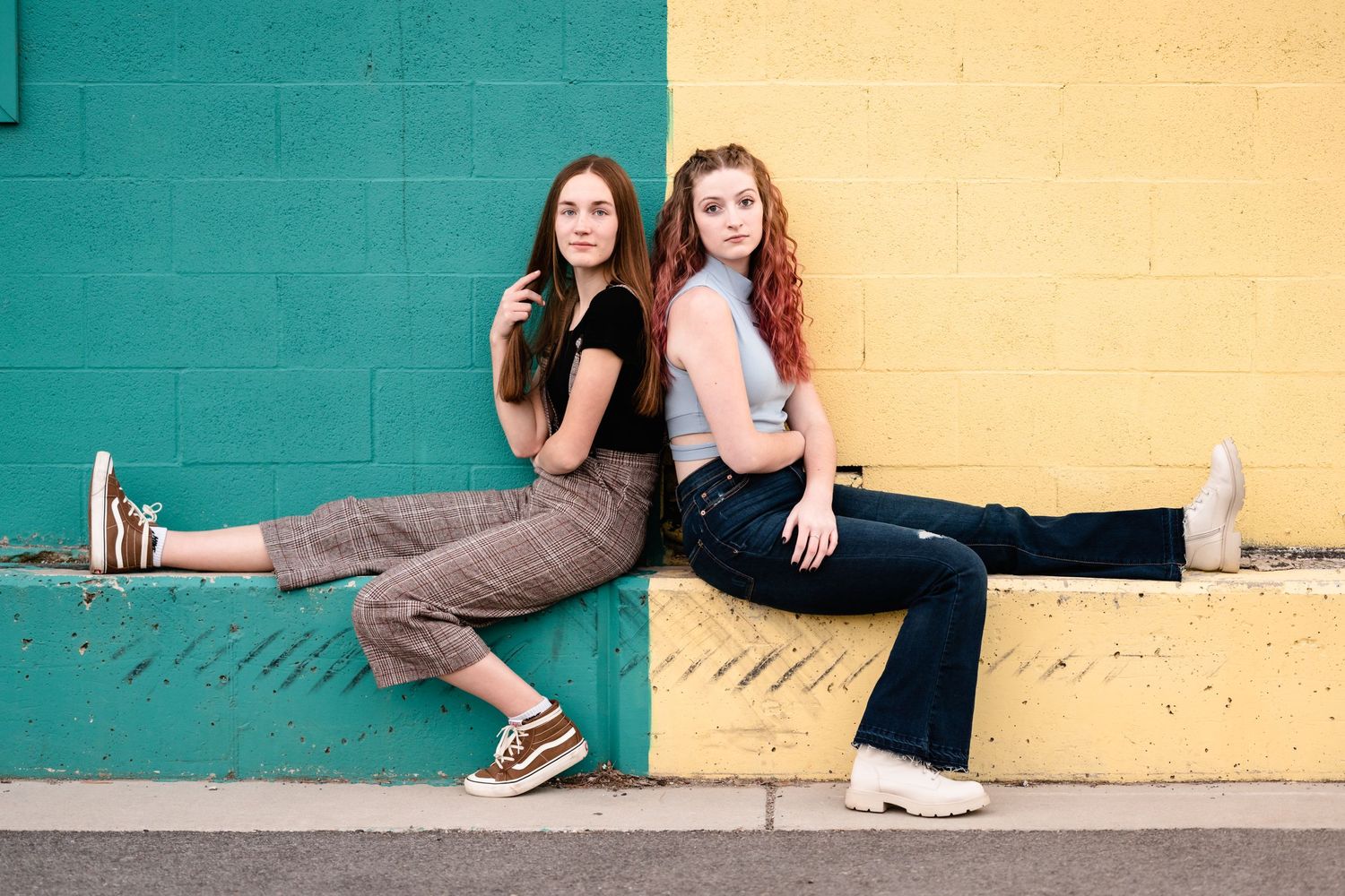 Two people in casual attire sit and lean against a colorful wall with teal and yellow sections.