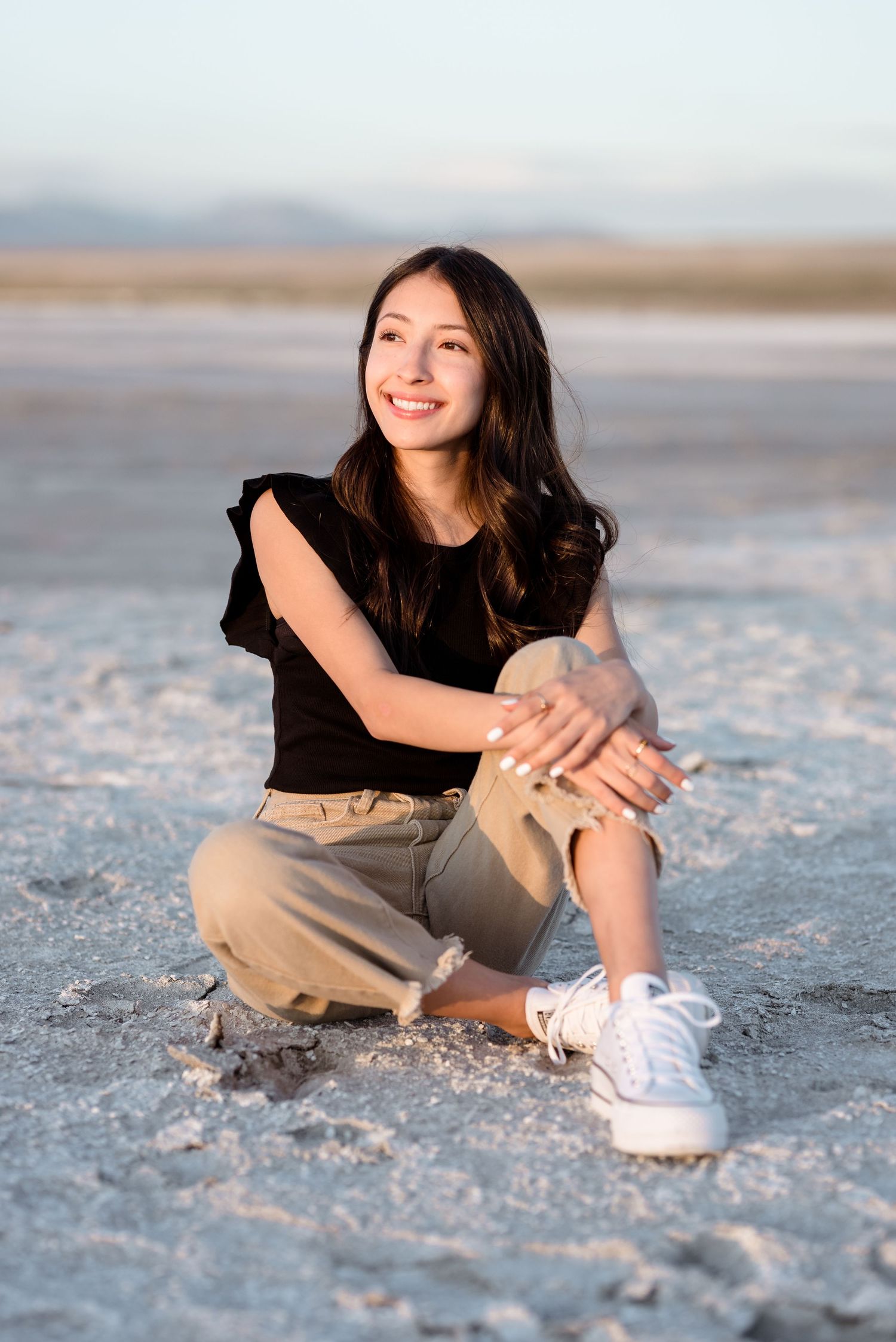 Person in black shirt and khaki pants poses casually on salt flats at sunset.