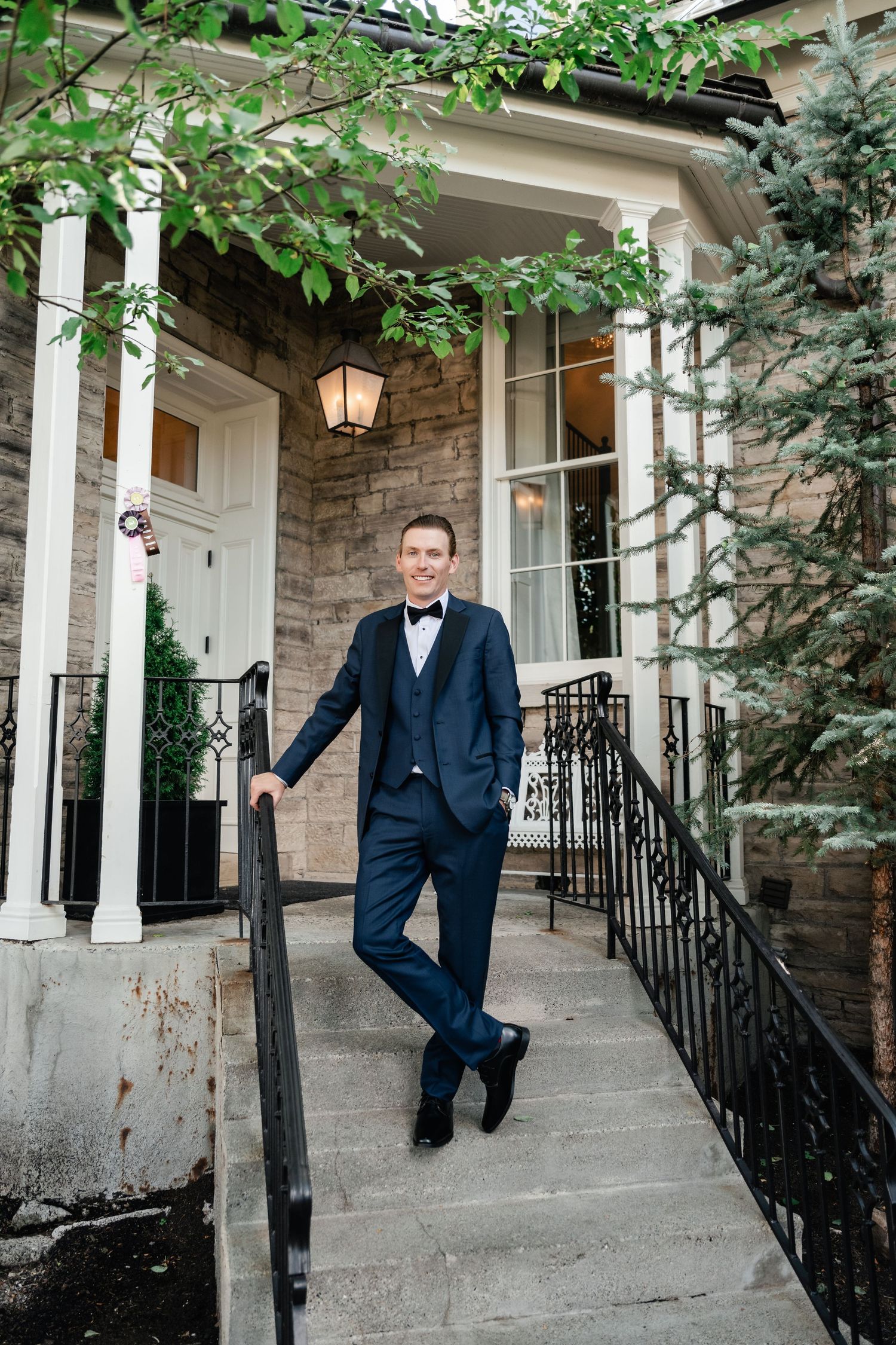 A smiling groom in a navy tuxedo with bow tie poses on stone steps outside the Washington School House Hotel during their intimate wedding.