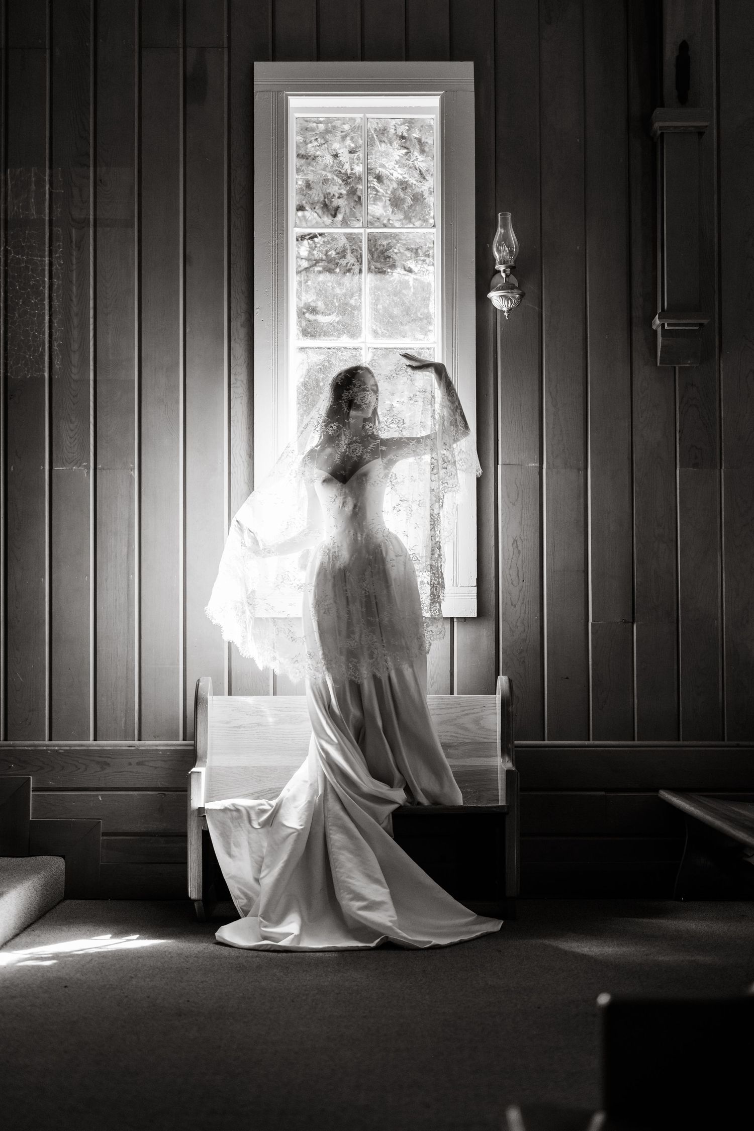 Black and white photo of a bride in a wedding dress and veil standing by a sunlit church window at the historic Yosemite chapel.