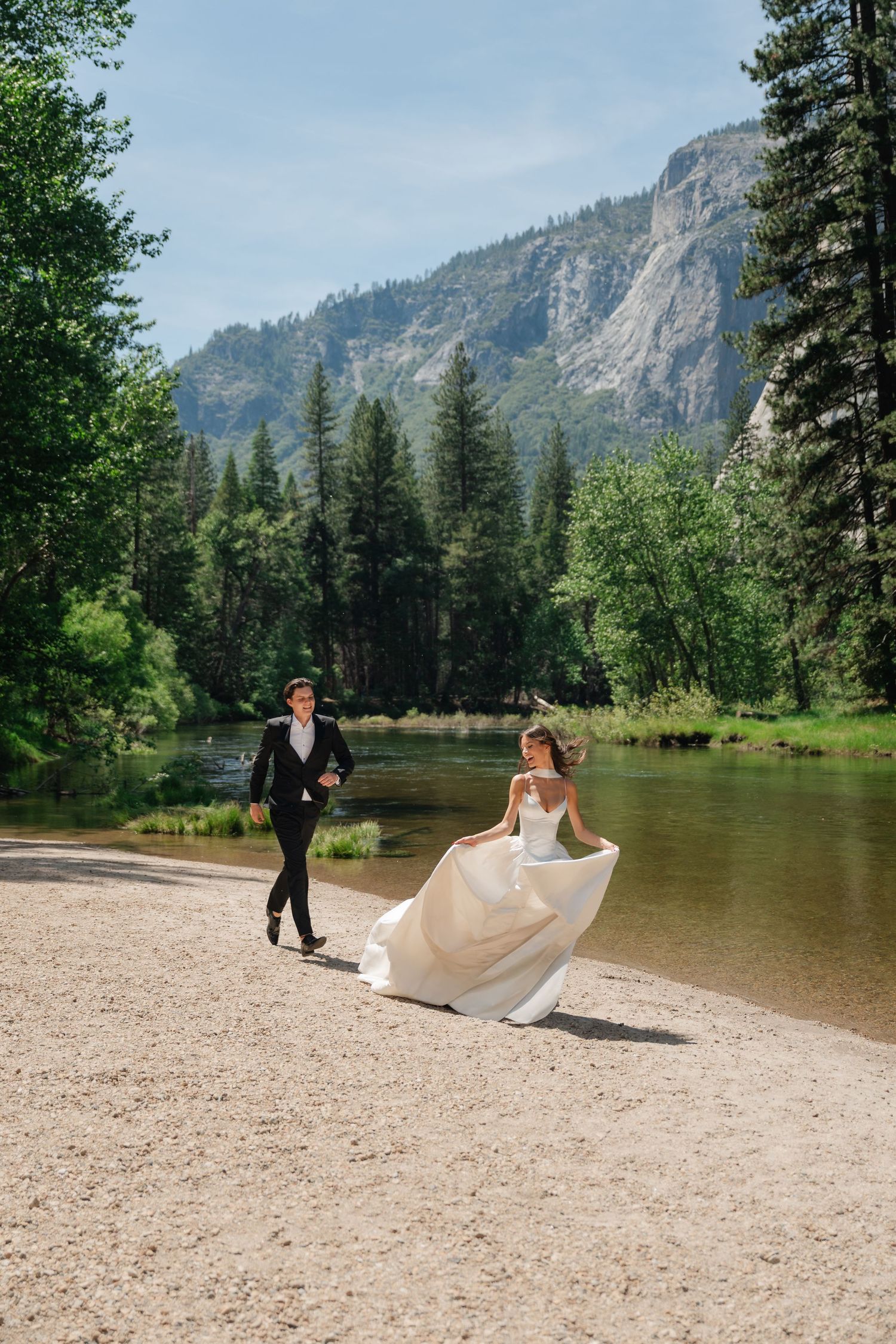 Bride and groom walking along a sandy riverbank with mountain and pine forest backdrop on their wedding day at Yosemite National Park.
