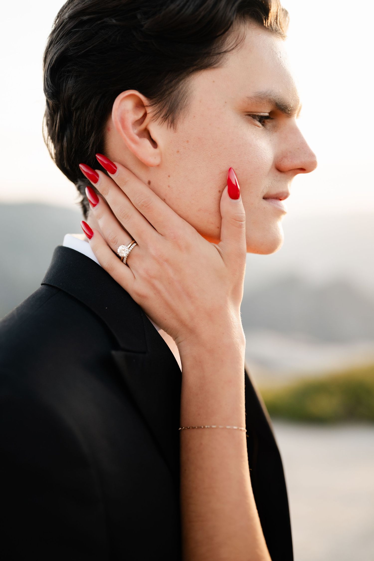 Close-up of bride with red nails and diamond ring gently touching grooms face outdoors.