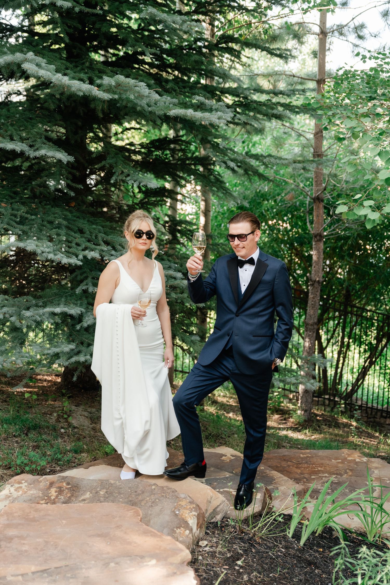 Wedding couple sits casually on garden rocks surrounded by evergreen trees and natural landscaping.