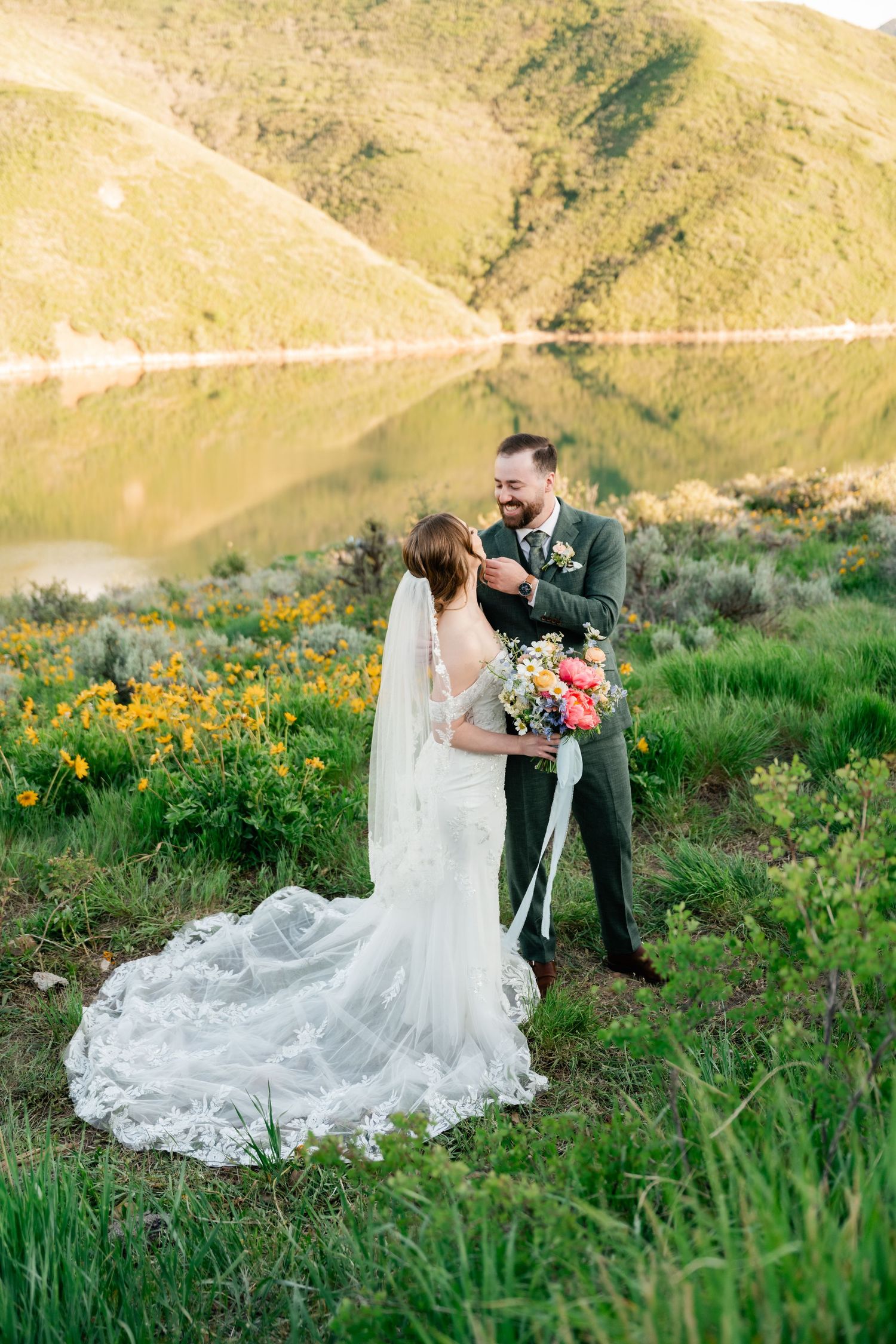 Bride in lace gown sits gracefully among yellow wildflowers with mountains in background.