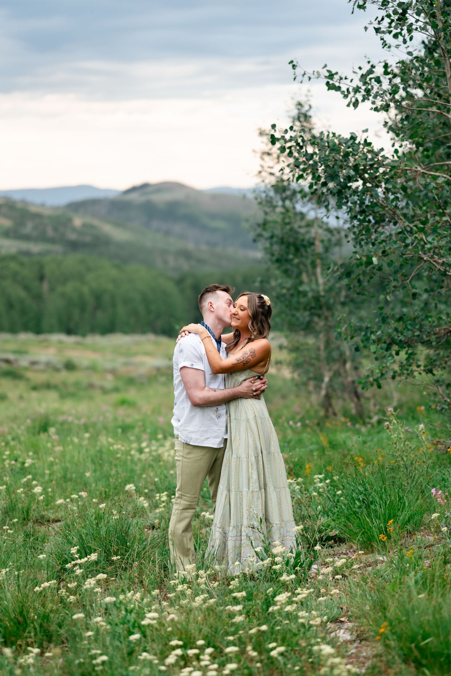 A couple embraces in a mountain meadow filled with wildflowers and distant peaks.