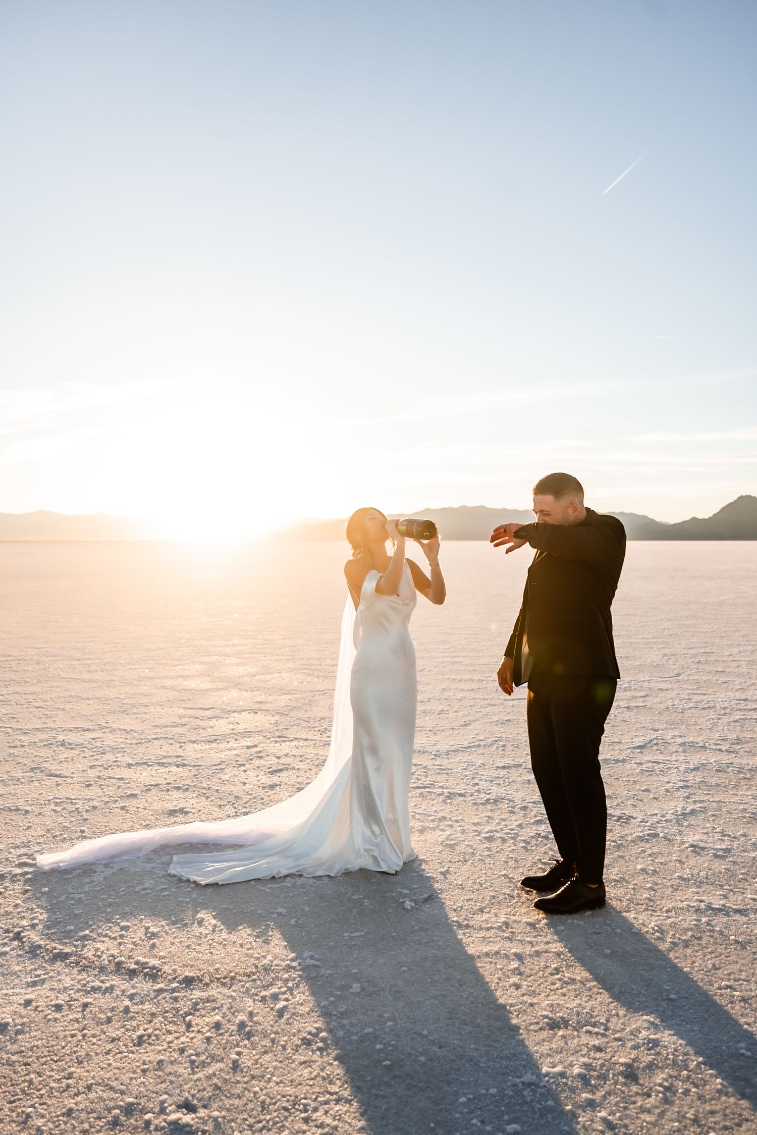 A couple in wedding attire playfully takes photos on salt flats at golden hour.