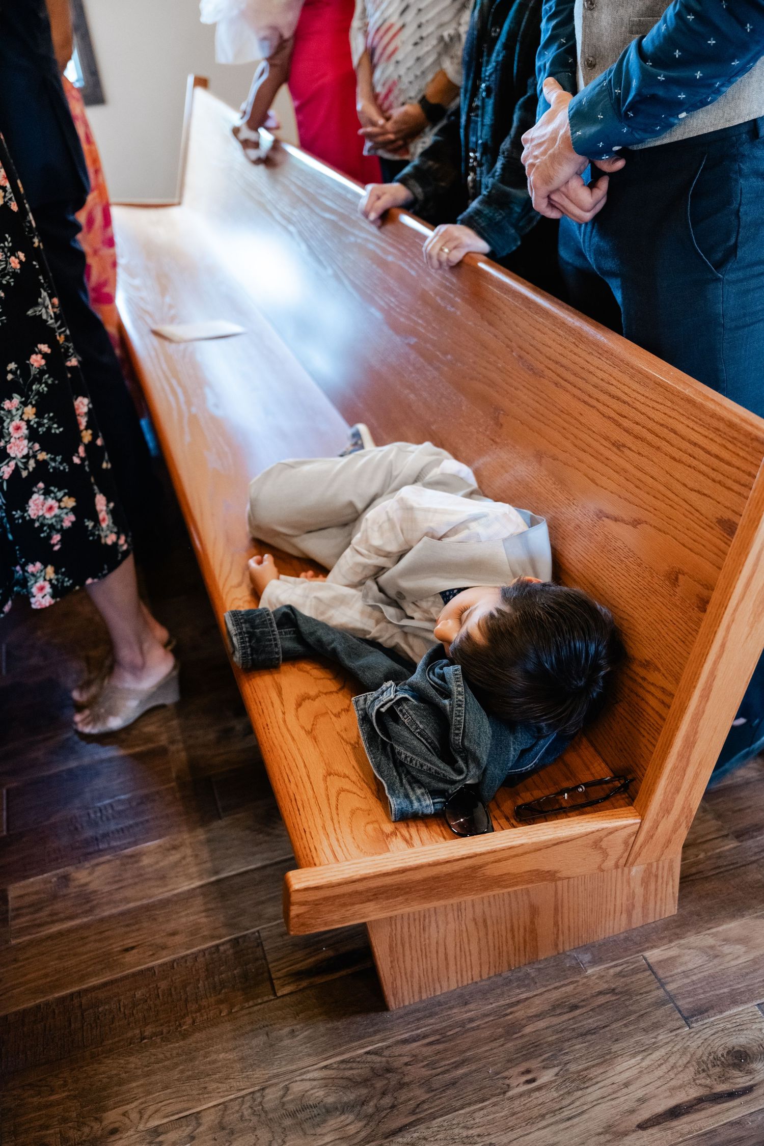 Young child lying down asleep on wooden church pew during ceremony.