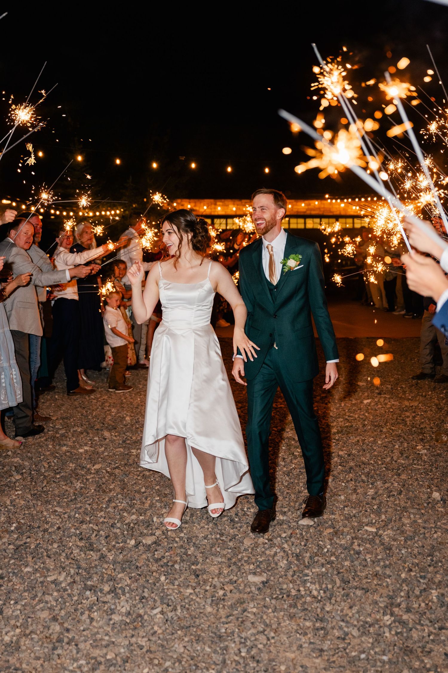 Wedding couple exits through sparklers at night under string lights on outdoor patio.