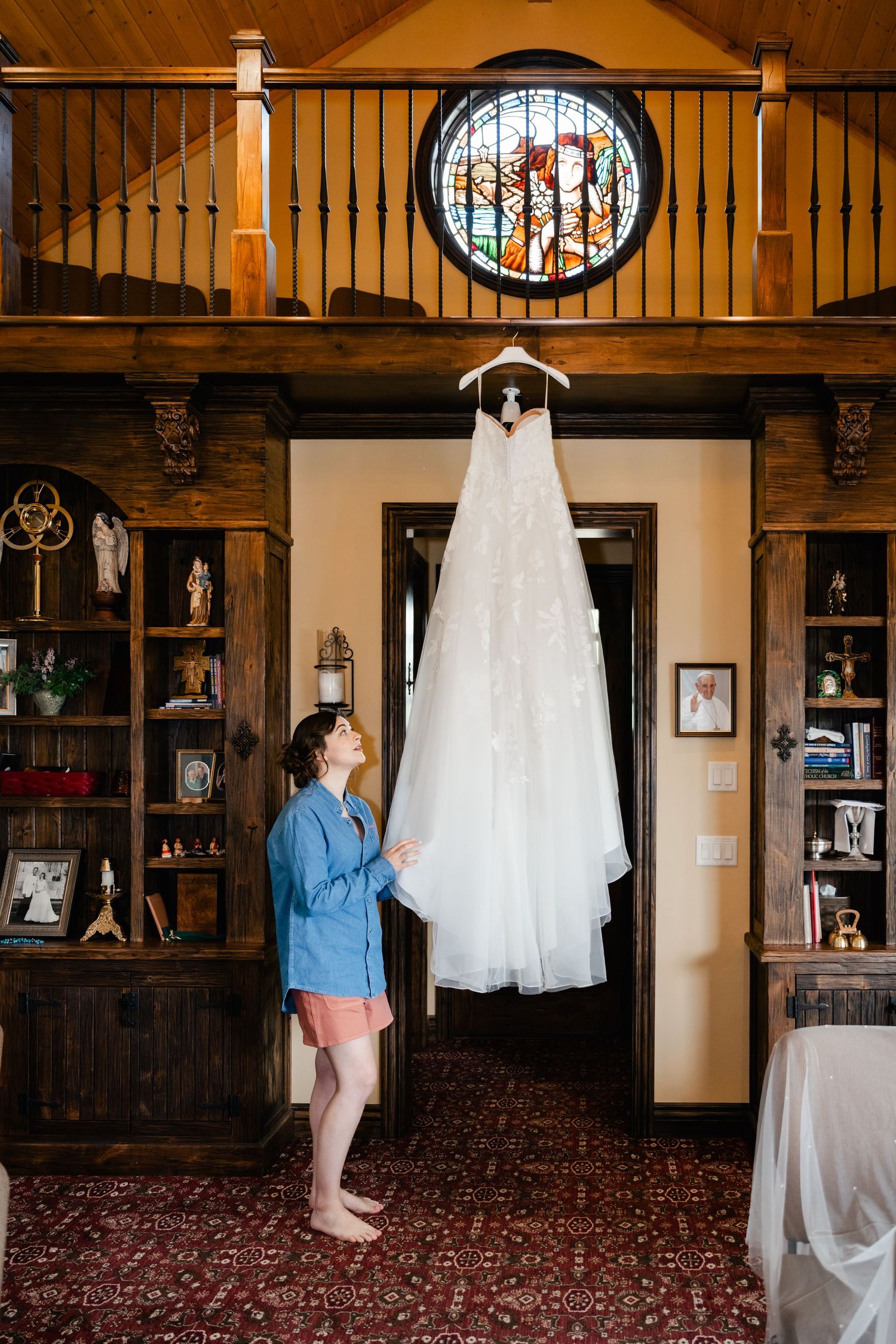 White wedding dress hanging in rustic wooden room with stained glass window.