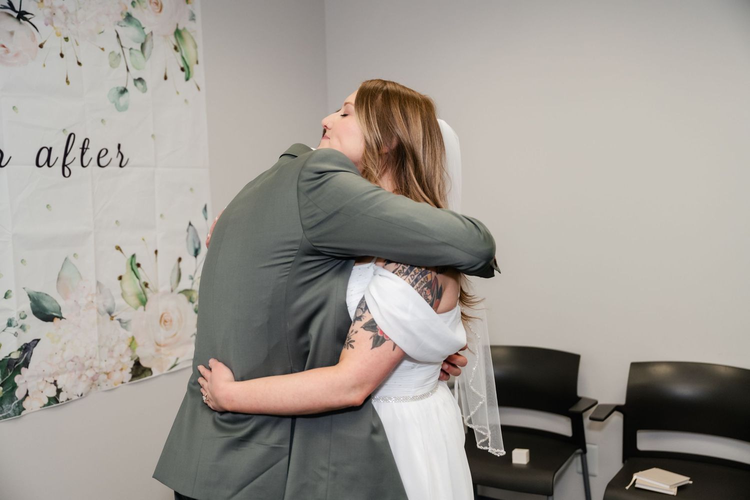Two people embrace in a heartfelt hug against a white wall with floral decorations.
