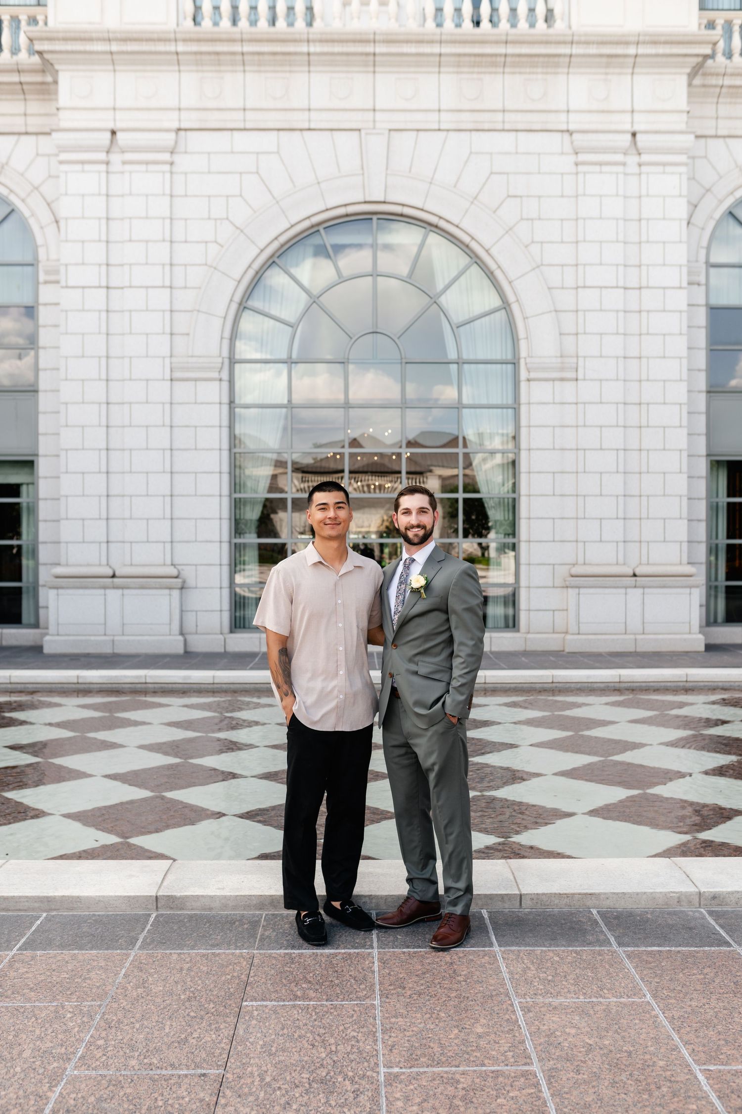 Two people stand in front of a grand arched entrance with white stone architecture and checkered floor tiles.