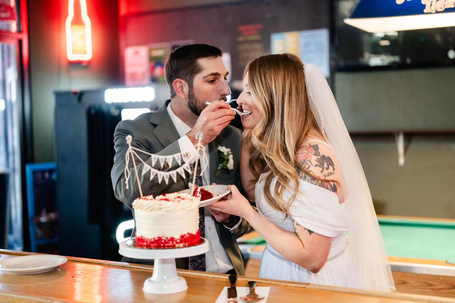 Newlyweds share cake and laughter during their reception at a casual bar venue.