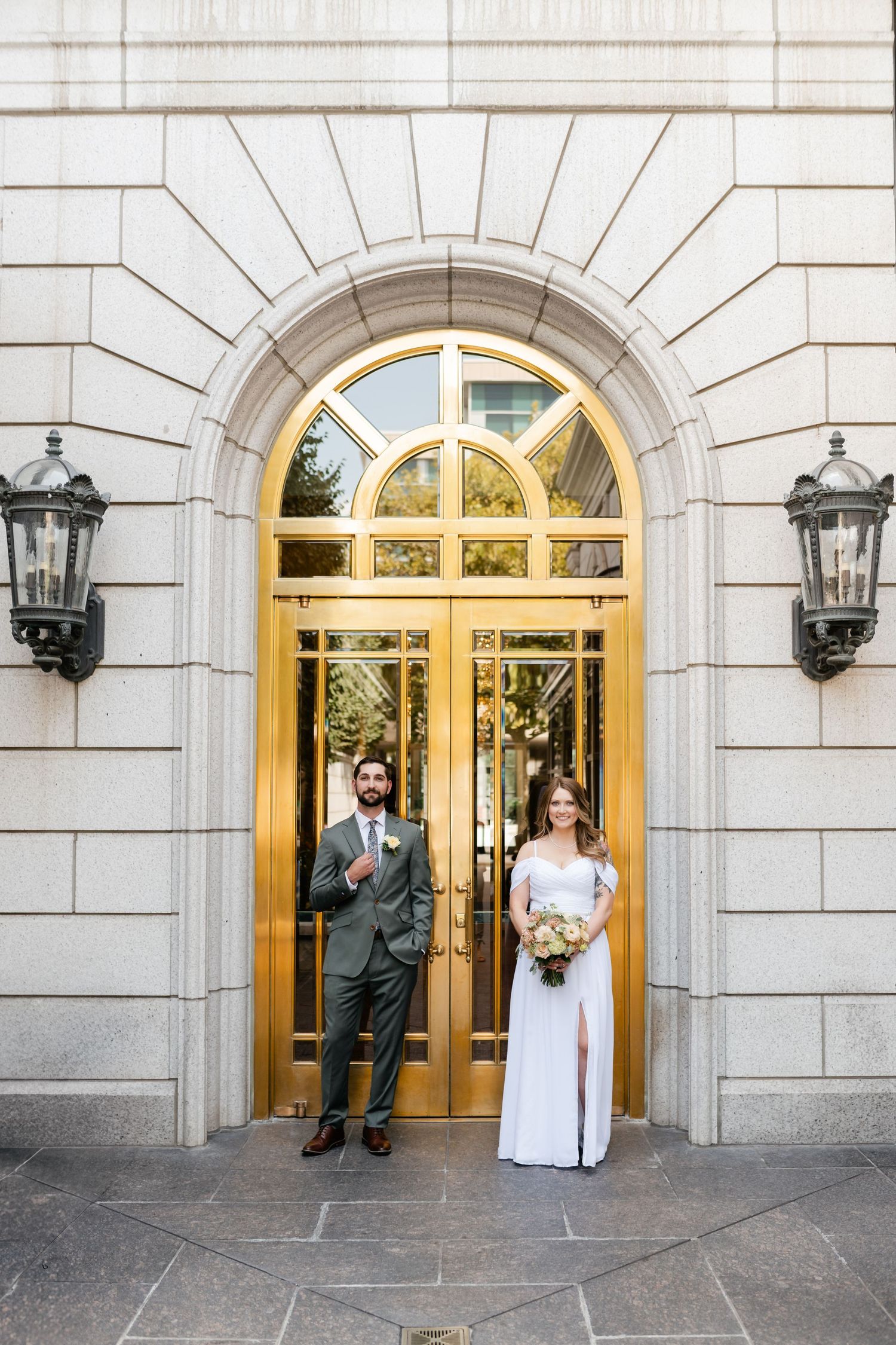 A bride and groom pose in front of elegant gold-trimmed doorways with stone architecture and wall-mounted lanterns.