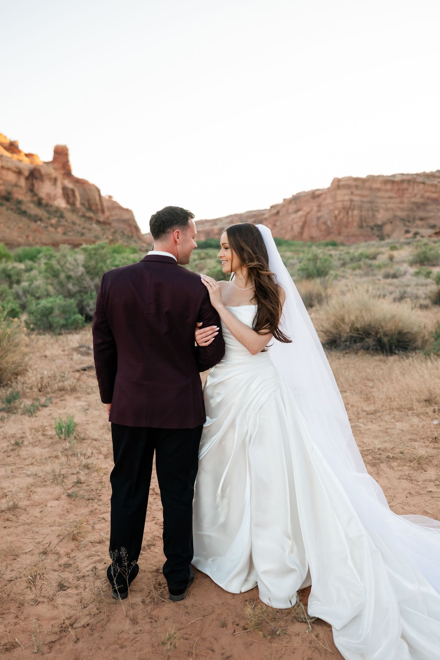 Bride and groom walk hand-in-hand through red rock desert landscape, bride wearing flowing white gown with long veil.