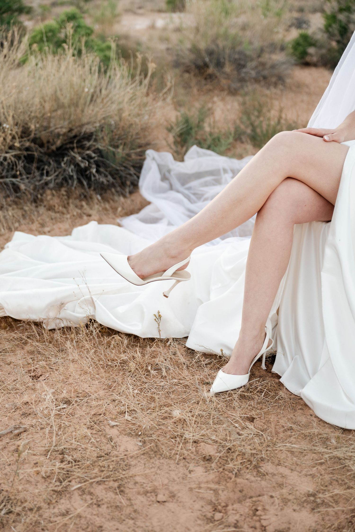 Close-up of bride's legs in white high heel shoes with flowing wedding dress train on desert ground.
