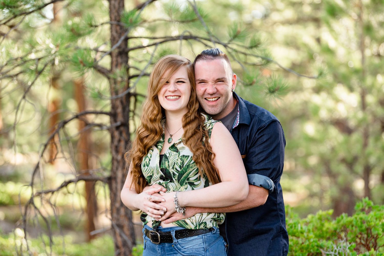 A couple poses together in a forest setting with natural greenery and sunlight filtering through the trees.