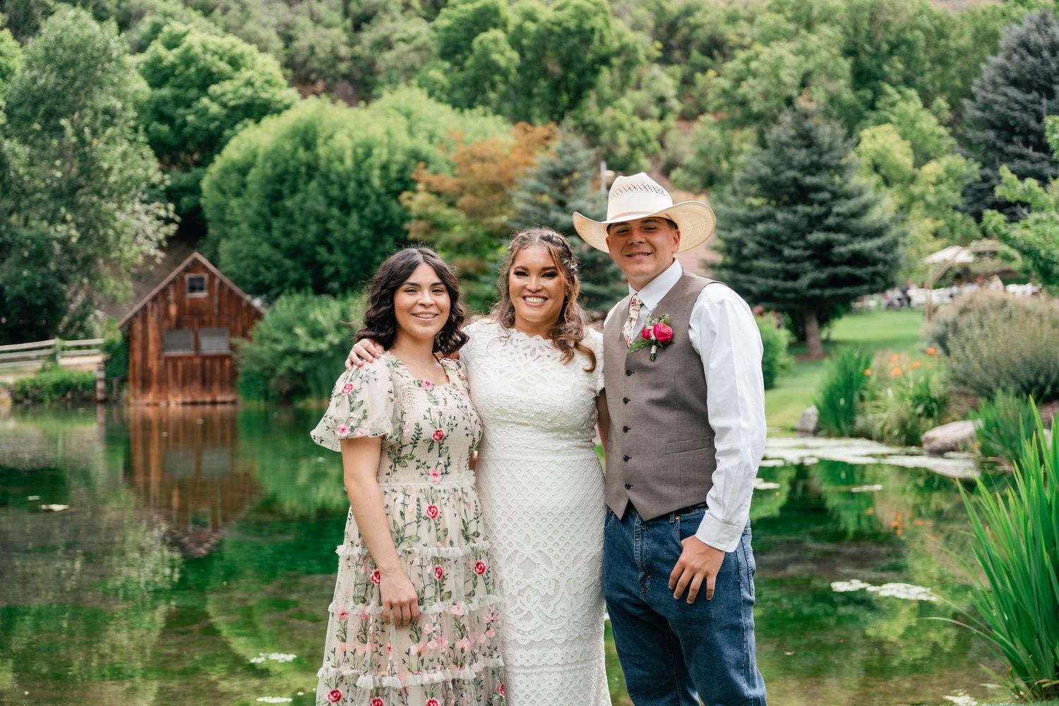 A family poses together near a serene pond surrounded by lush greenery and trees on a sunny day. Conrad Ranch, Utah Wedding Venue