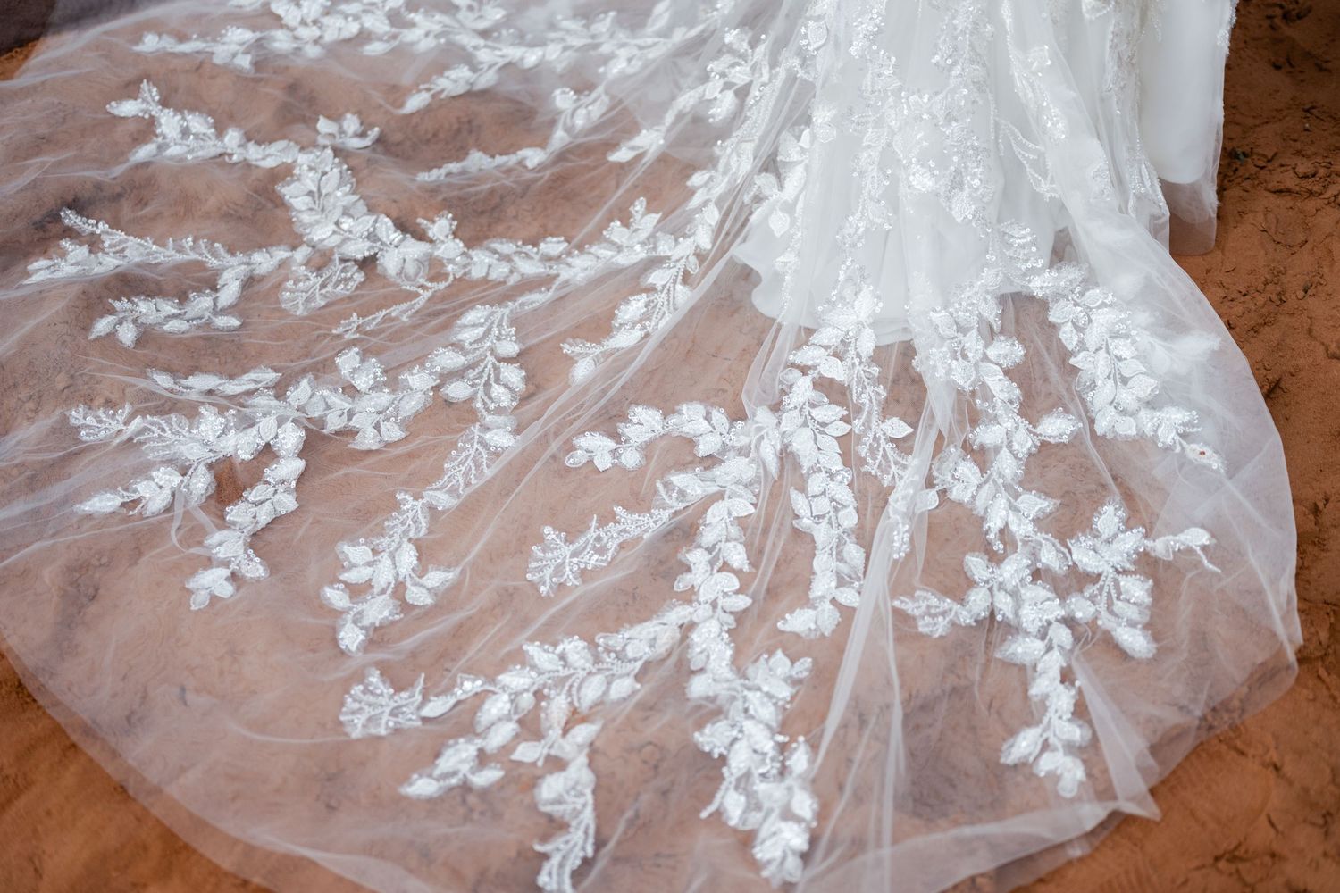 Intricate white lace details and floral patterns on a wedding dress train spread across desert ground.