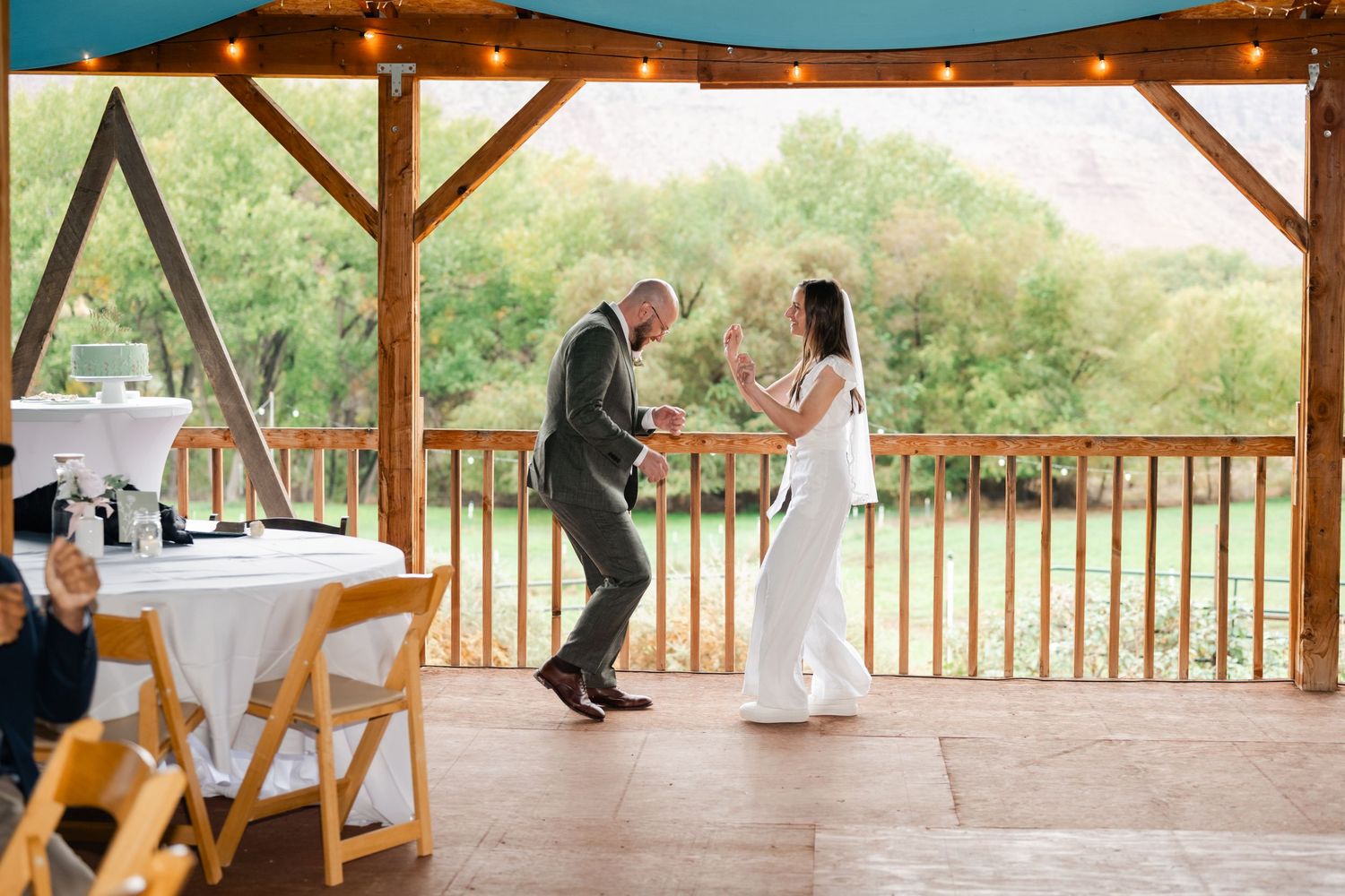 A couple dancing together on a wooden deck pavilion with string lights and mountain views.